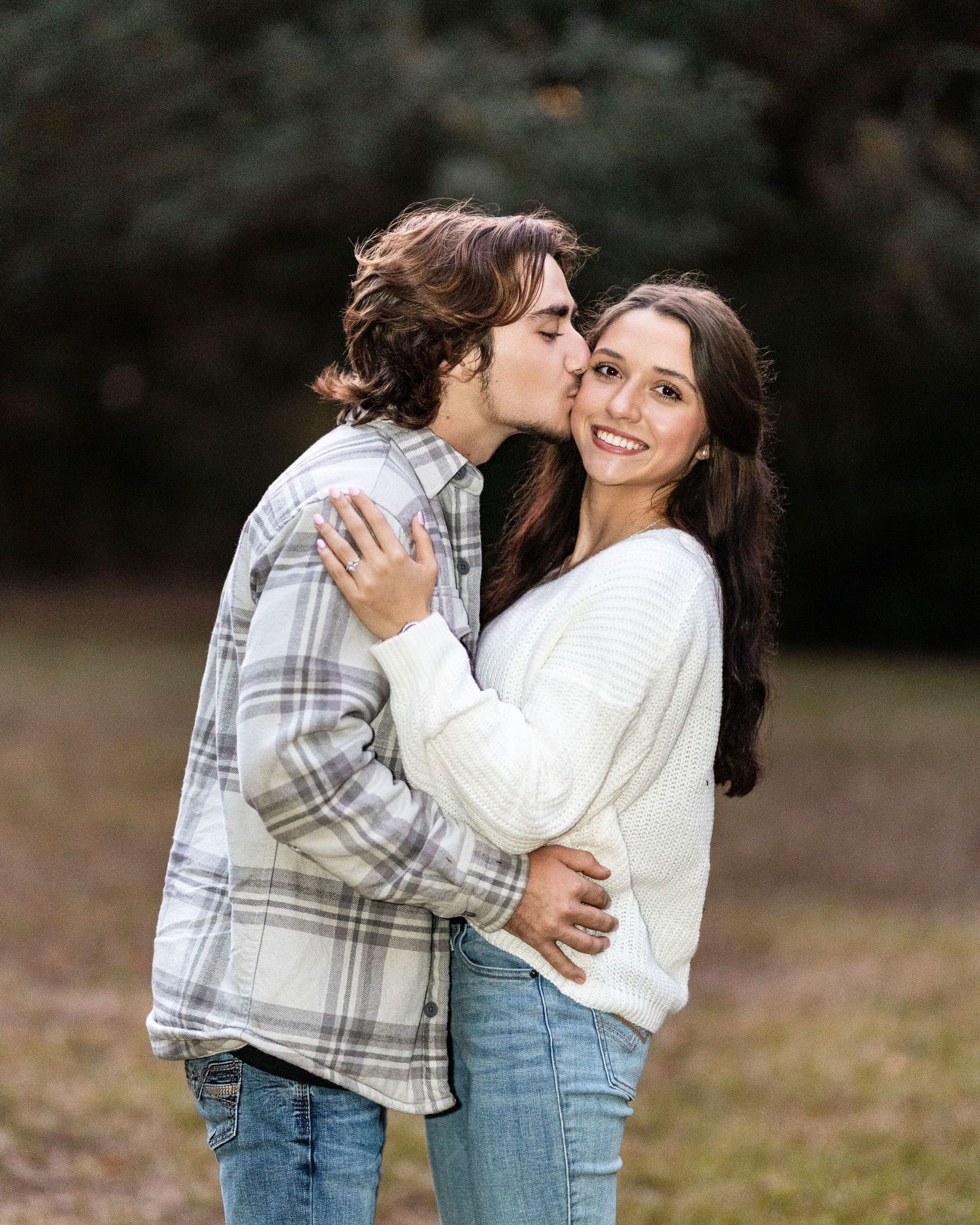 A romantic outdoor portrait of a couple embracing, with one wearing a plaid shirt and the other in a white sweater.
