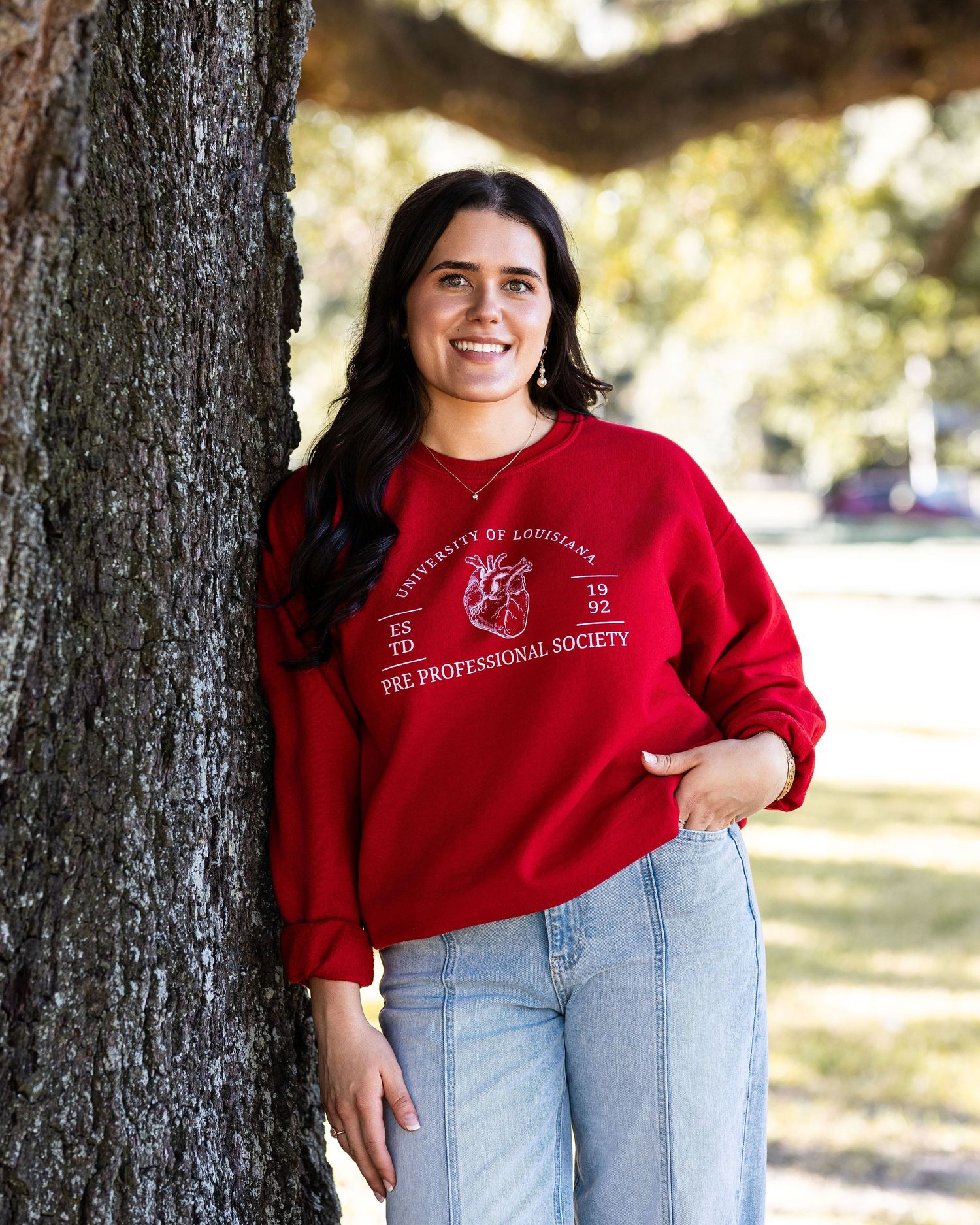 A person wearing a red sweatshirt and light jeans sits comfortably against a large tree in a sunny park setting.