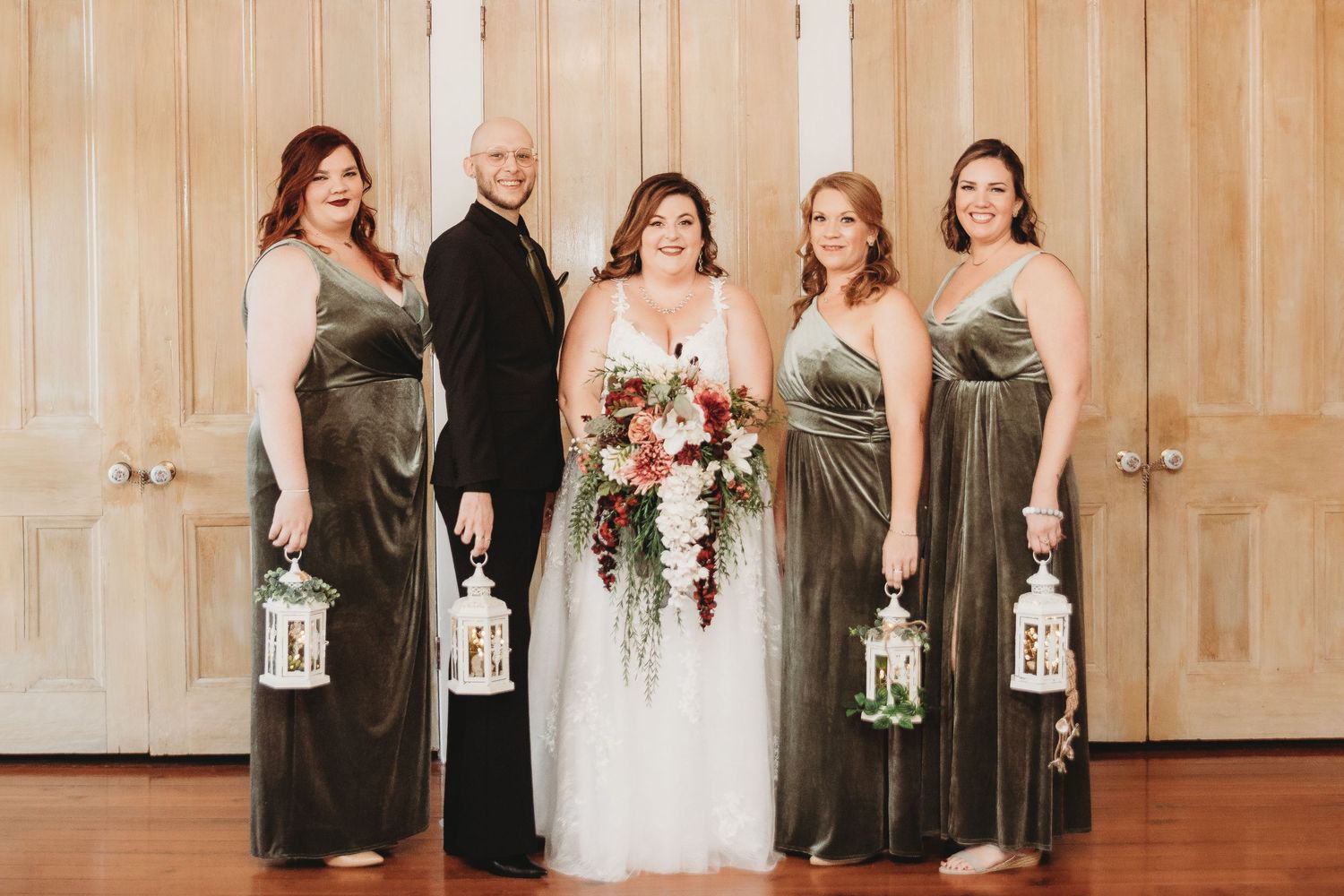 Wedding party poses with lanterns against wooden doors in elegant green dresses.