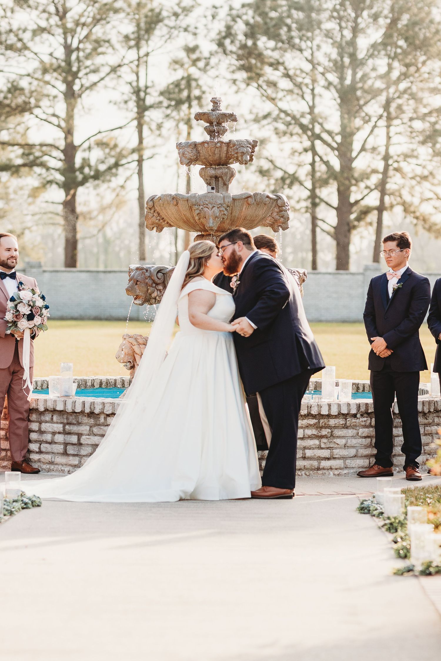 Elegant outdoor wedding ceremony with stone pillars and fountain surrounded by towering pine trees.