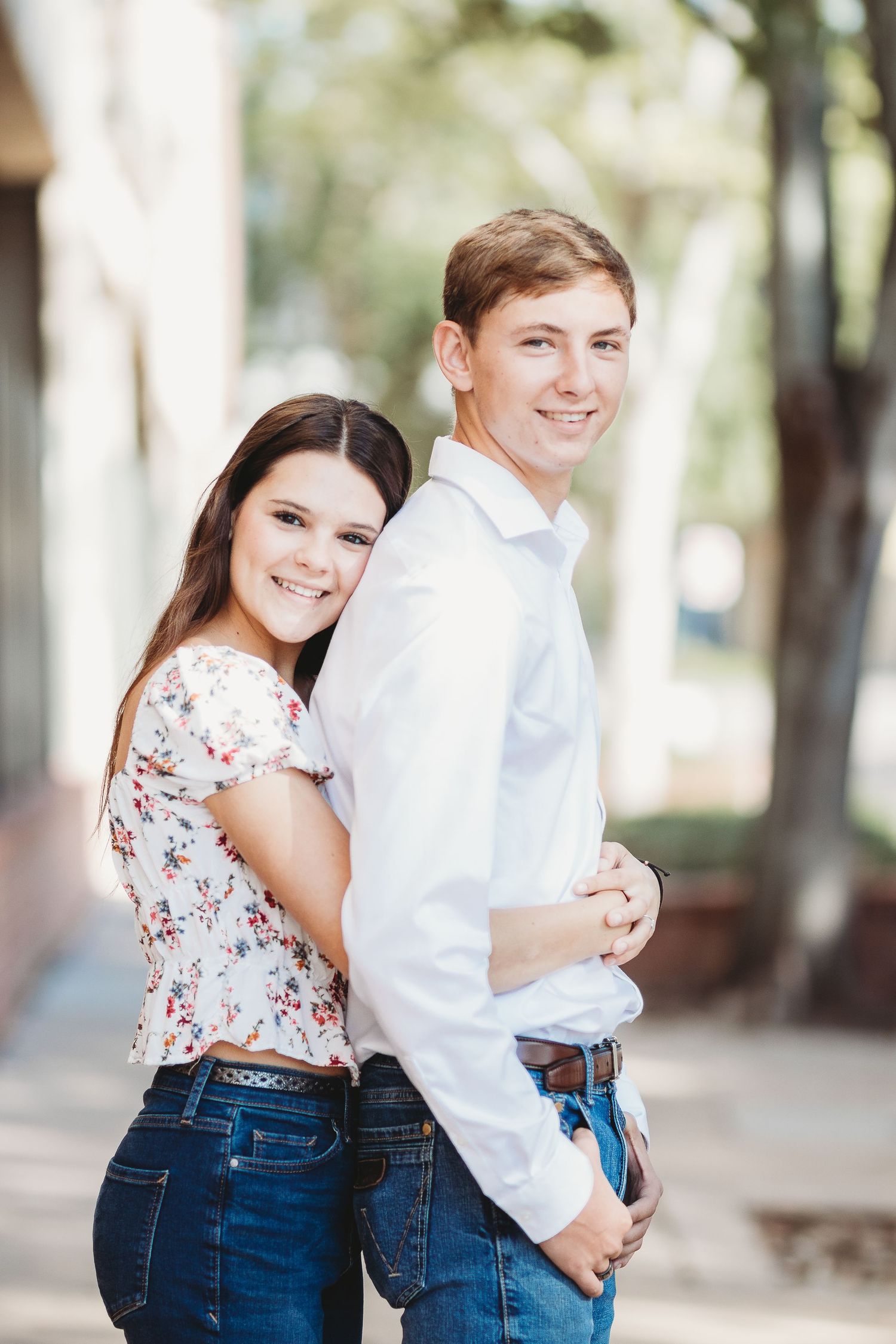 Two people in matching jeans and light tops embrace while sharing candid moments outdoors.