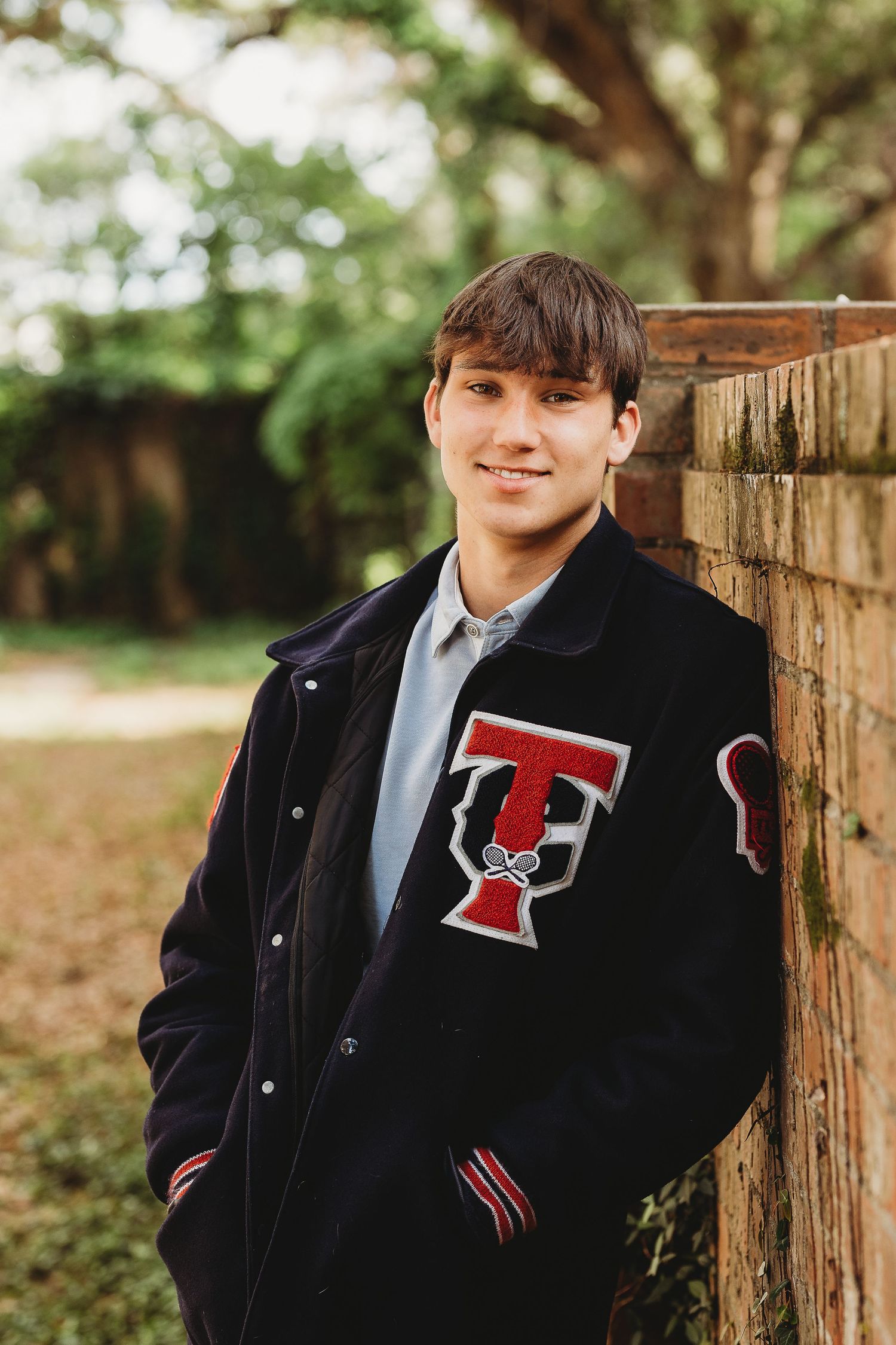 Student wearing black varsity jacket with red T emblem leans against brick wall outdoors.
