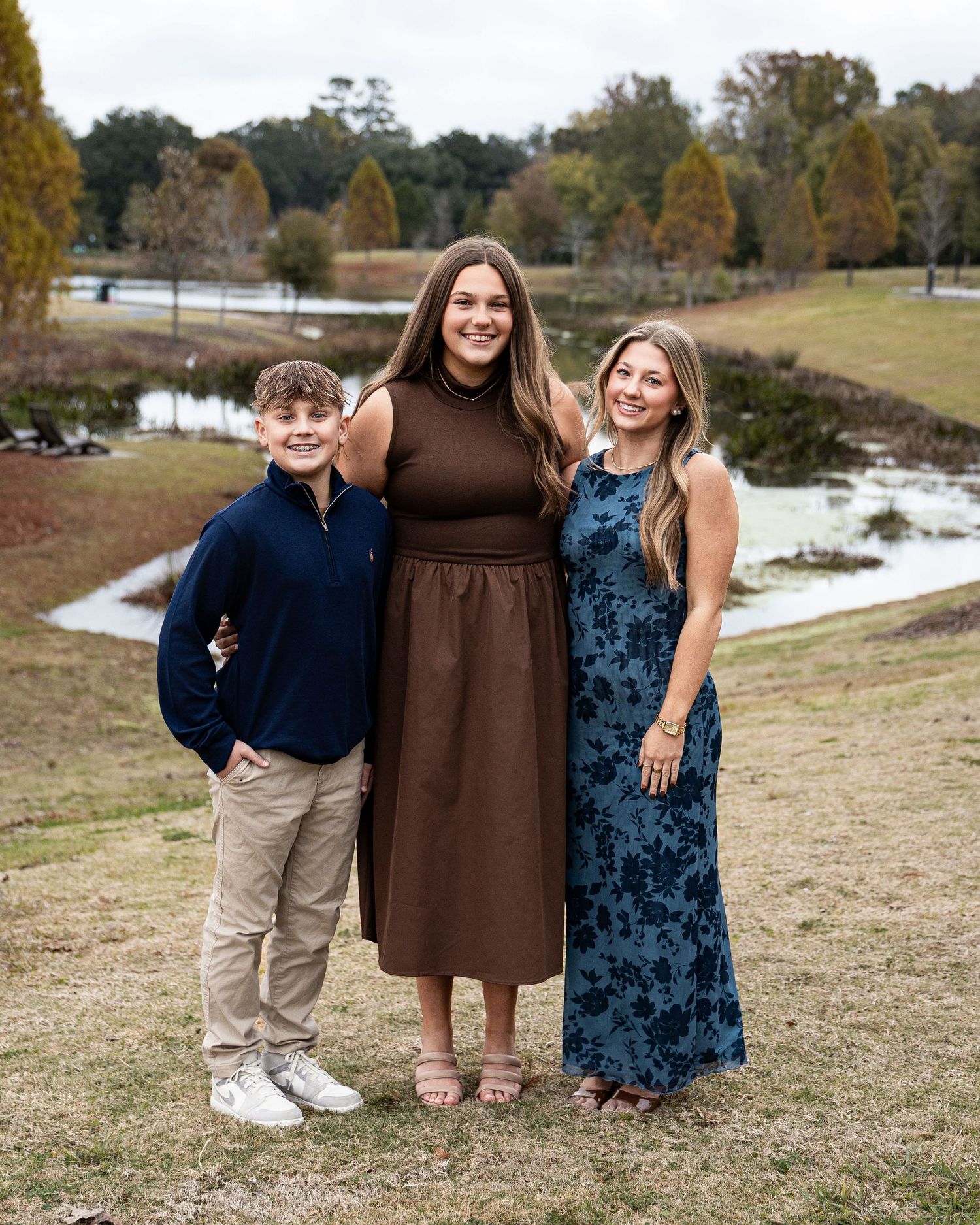 A group stands together outdoors on a grassy hill during autumn with trees and a lake in the background.