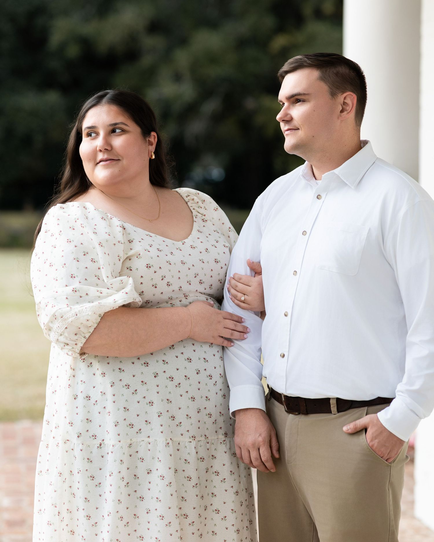 Romantic couple in white dress and shirt pose together against white pillars outdoors.