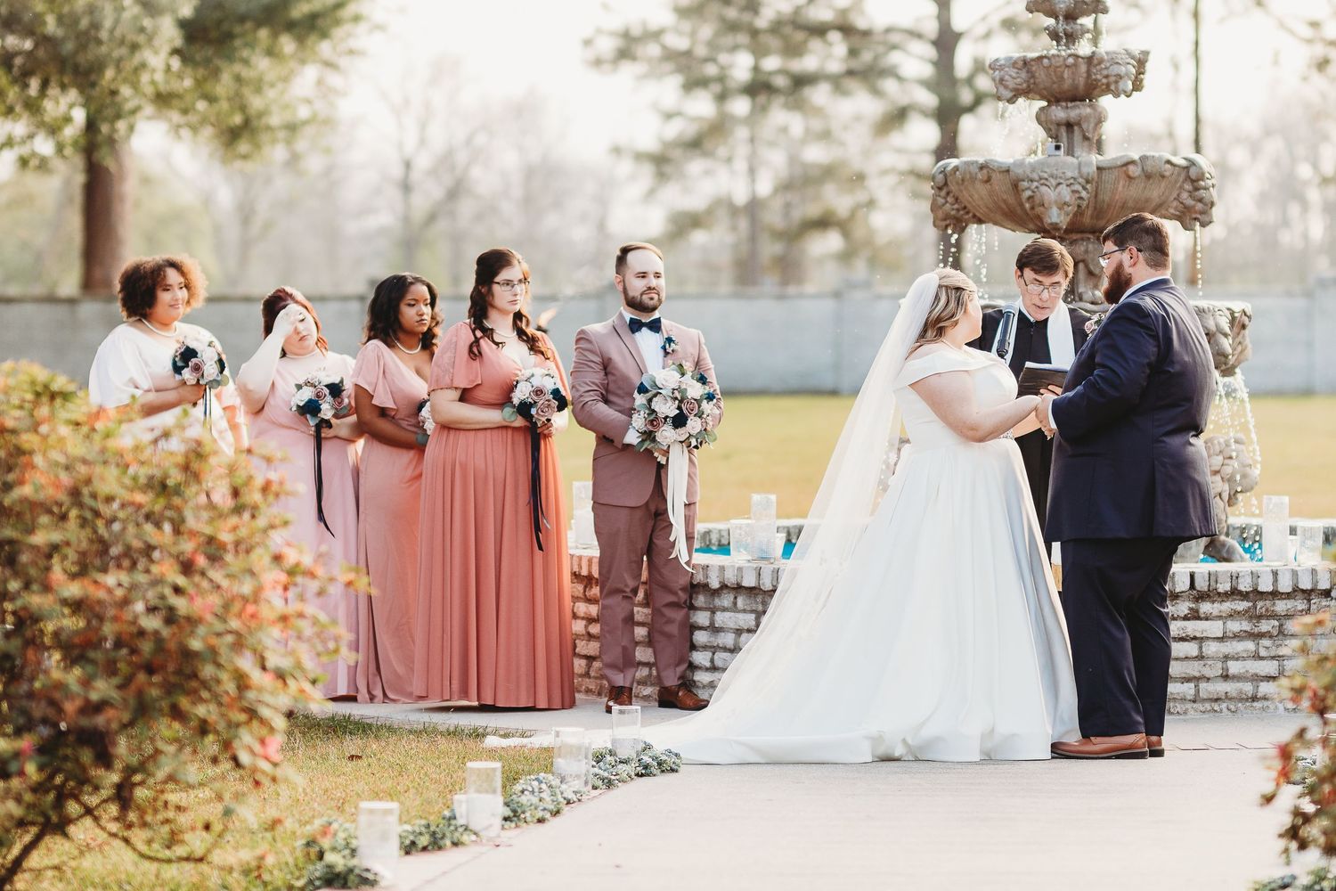 Wedding party members gather for outdoor photos near a decorative fountain.