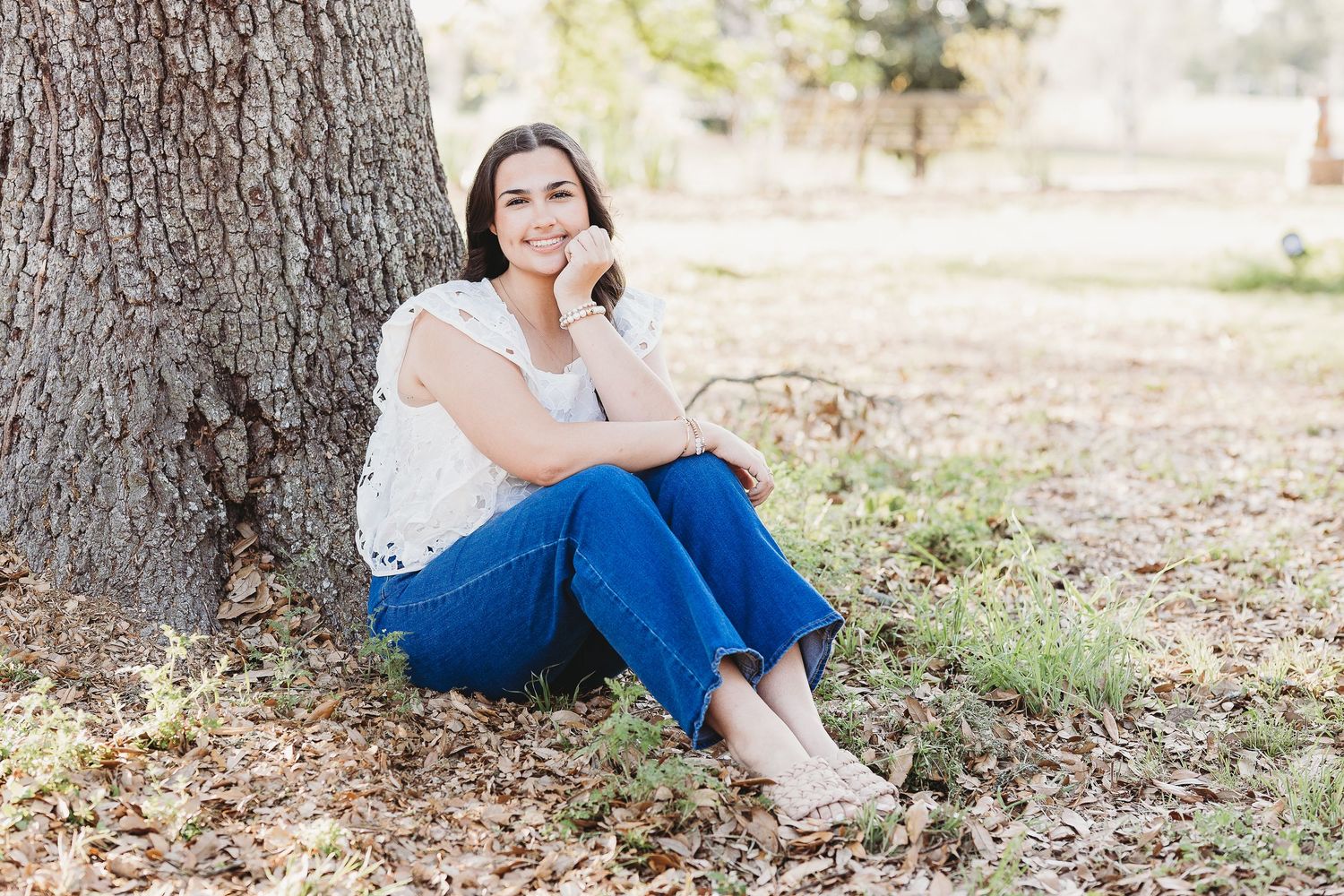 Casual outdoor portrait leaning against large tree trunk wearing white top and jeans.