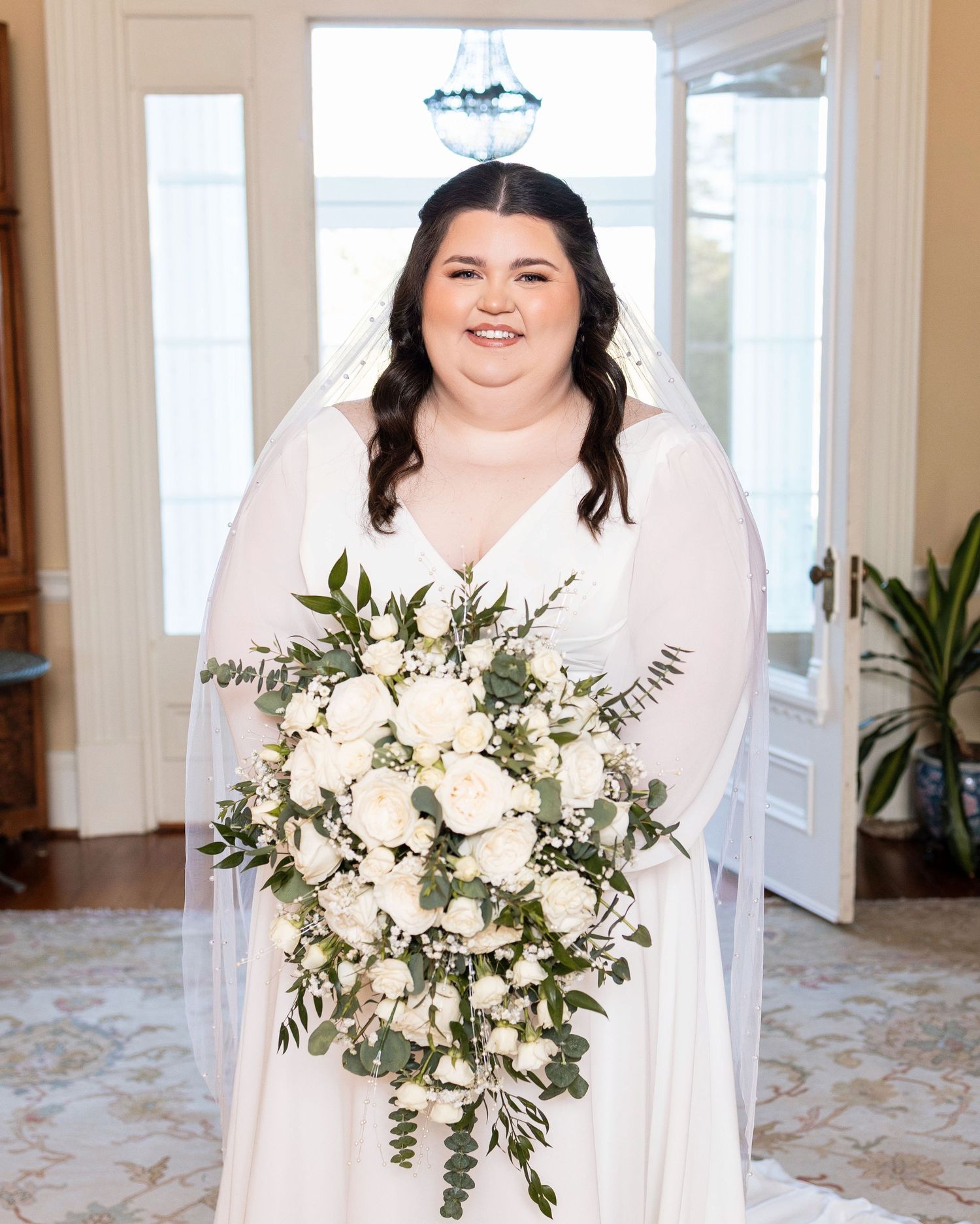 Bride in elegant white dress holding a lush bouquet of white flowers and eucalyptus in a bright indoor setting.