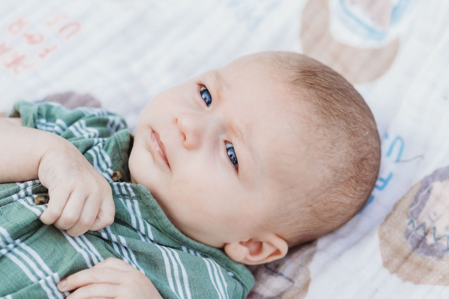 A baby in striped clothing lies on a patterned blanket looking up at the camera.