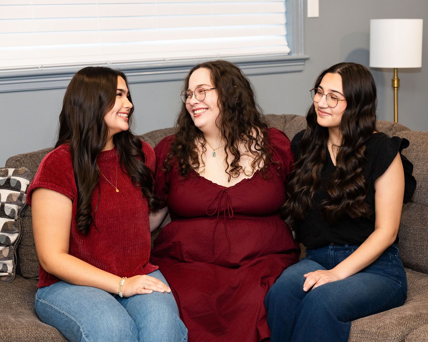 Three women in coordinating outfits sit together on a couch, sharing laughter and smiles in a casual indoor setting.