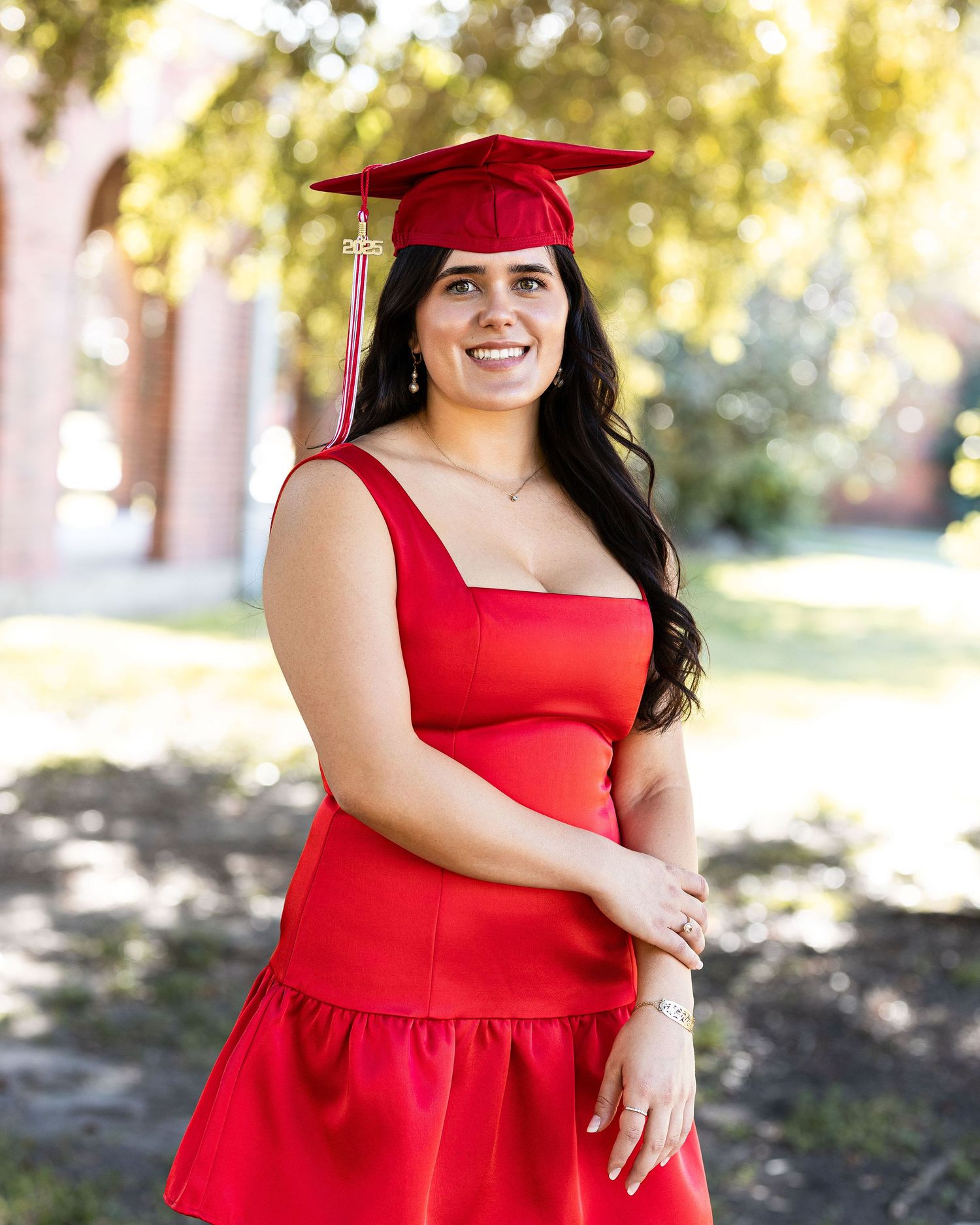 Graduate in a red cap and matching red dress poses outdoors on a sunny day with trees in the background.
