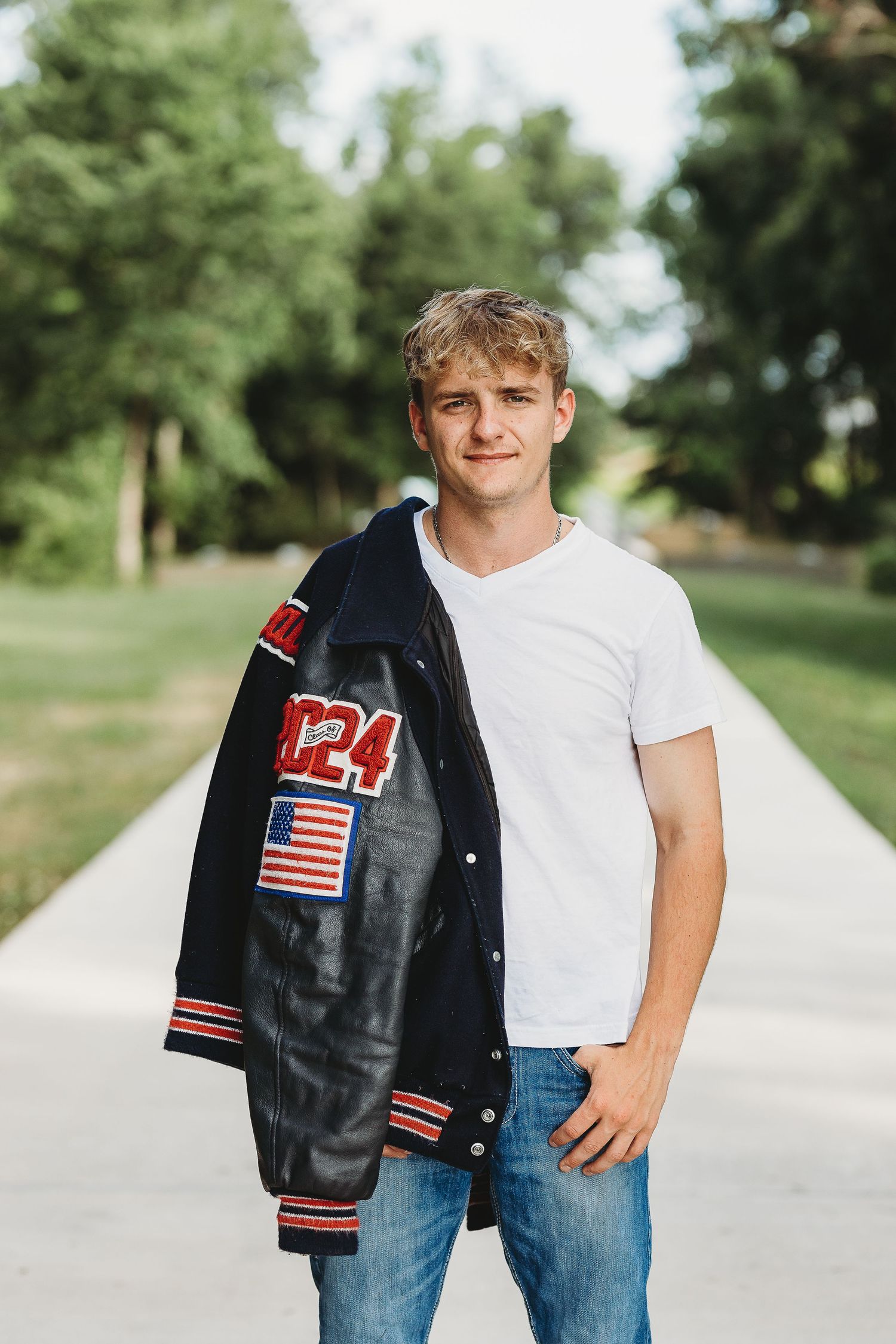 Student wearing varsity letterman jacket with American flag patch poses outdoors on path.