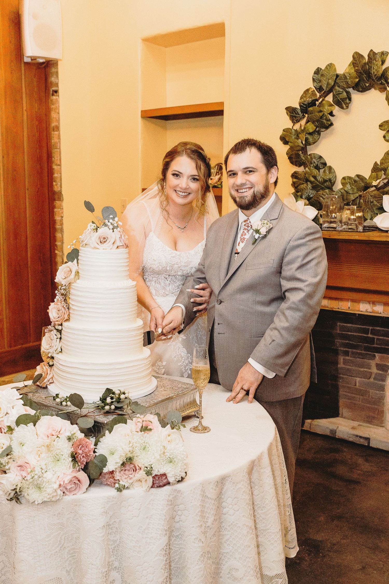 Couple cuts their white tiered wedding cake adorned with blush roses and greenery.