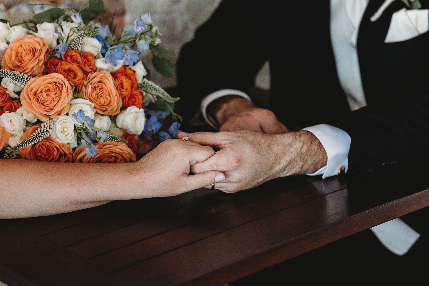 Wedding couple's hands with orange and blue floral bouquet on wooden table.