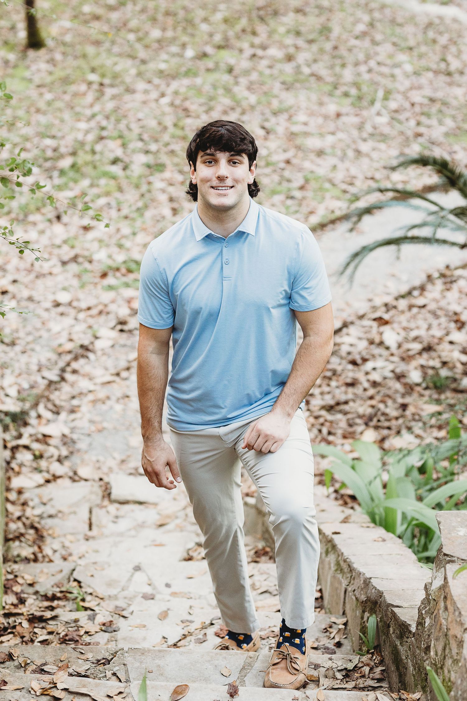 Person in light blue polo and khakis sits casually on tree stump in wooded area.