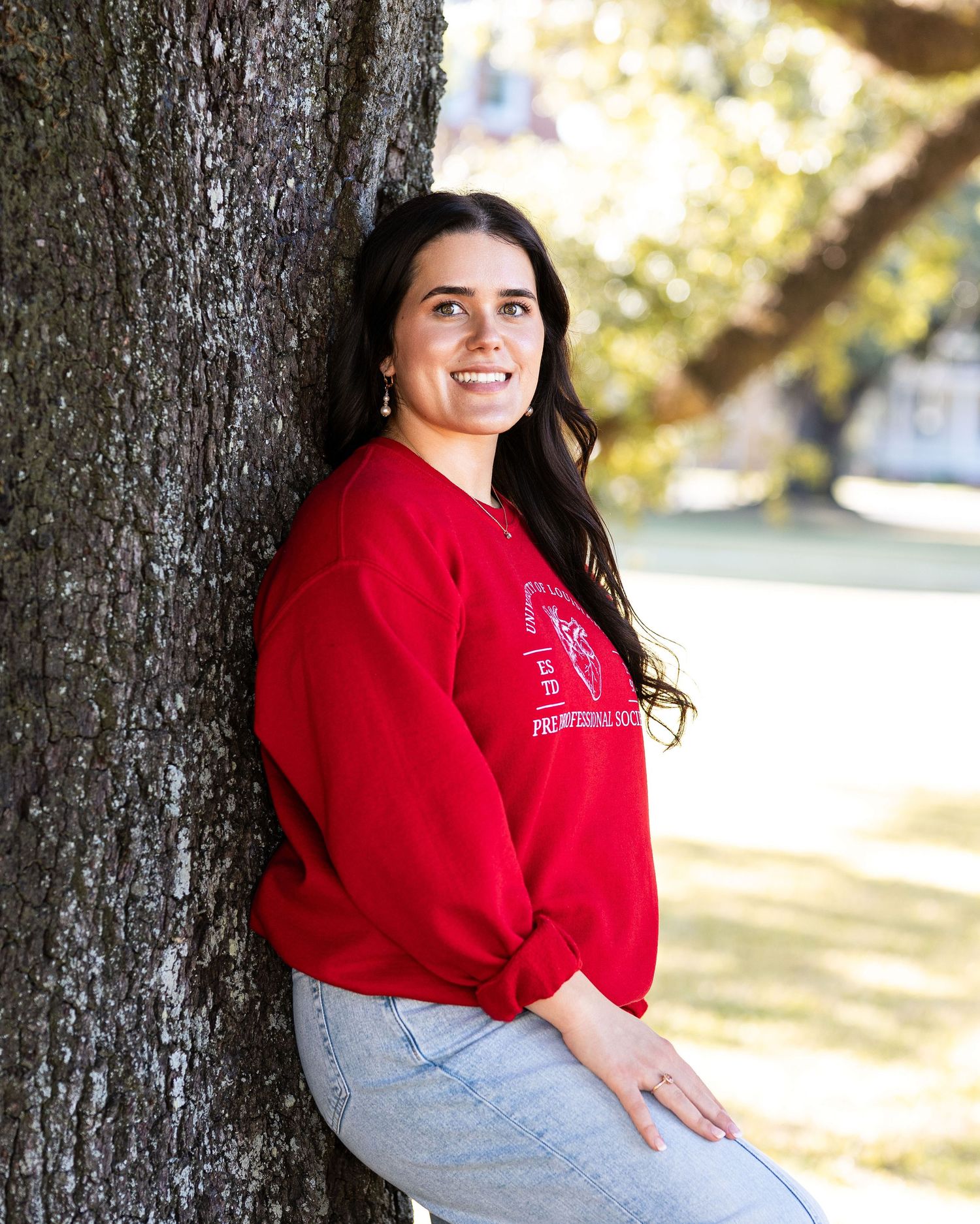 A person in a red sweater and jeans poses casually against a tree trunk in an outdoor setting.