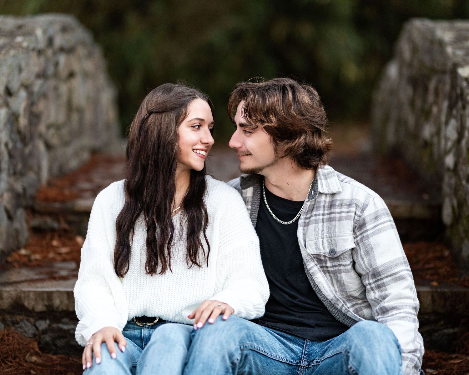 A couple sits together on fallen logs in a wooded forest setting during an autumn photoshoot.