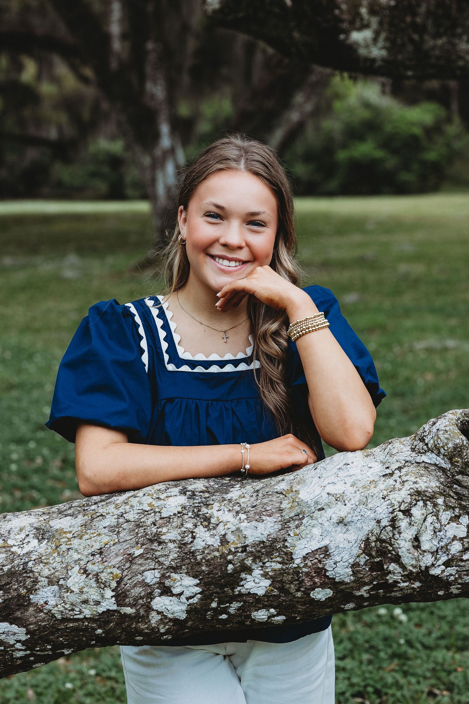 Someone in a navy blue dress poses playfully against a moss-covered tree branch outdoors.