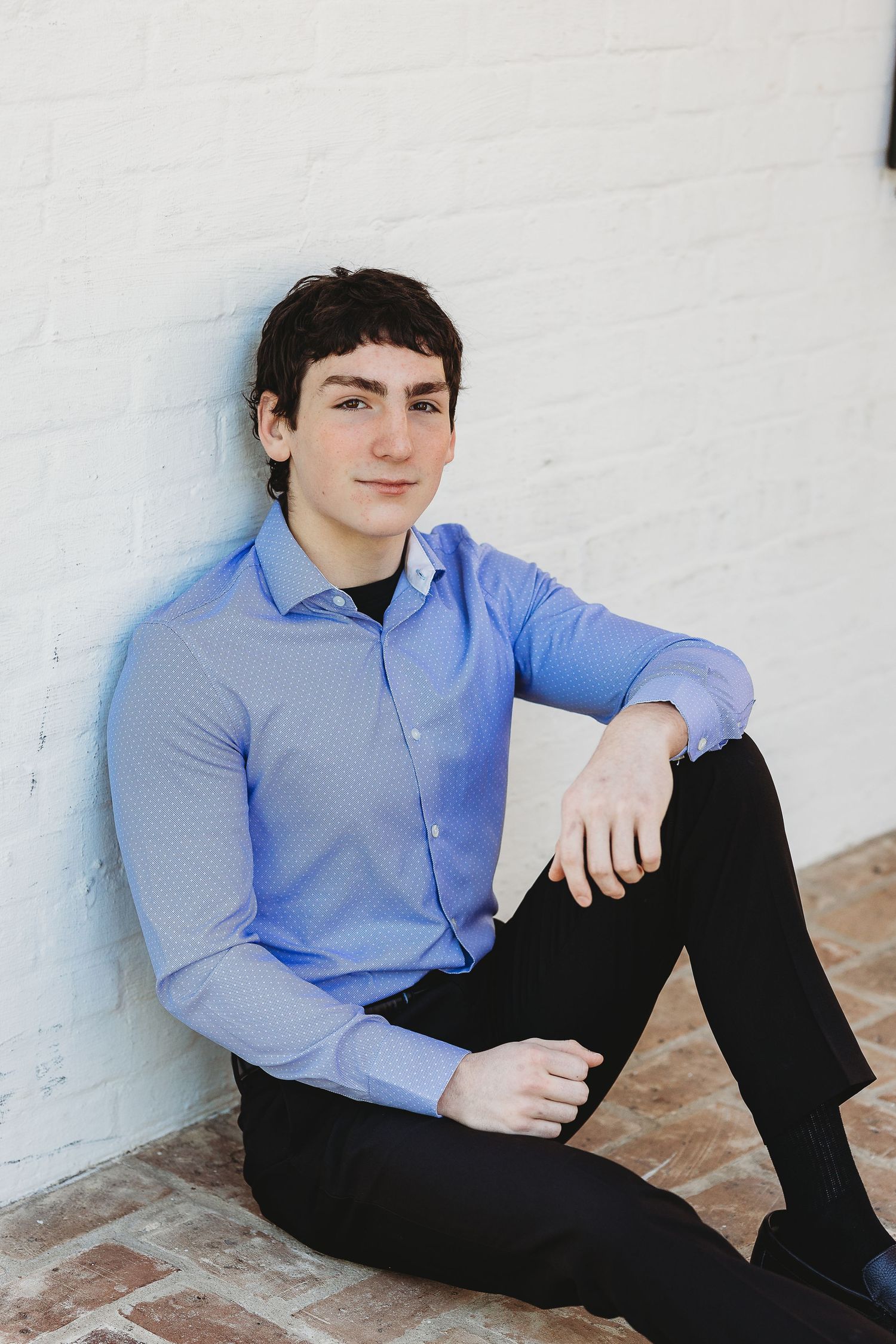 A casual portrait of someone in business attire sitting against a white wall.