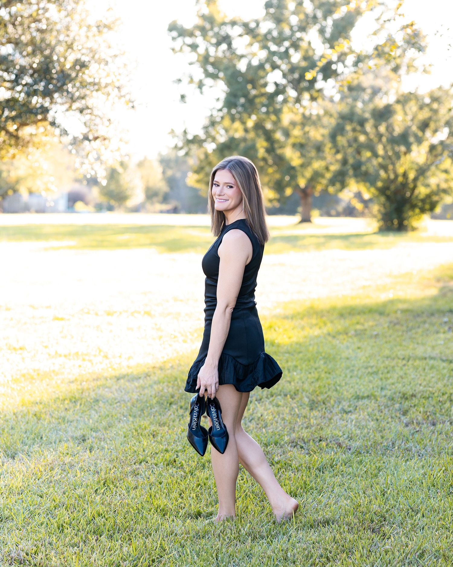 A person wearing a black dress spins playfully on a grassy lawn during golden hour.