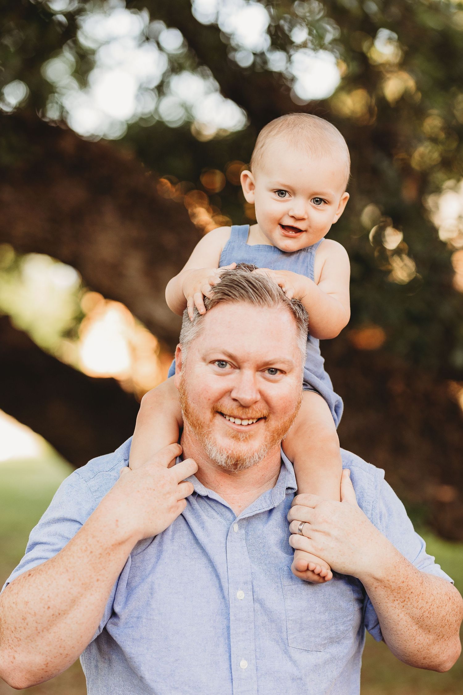 A playful outdoor family portrait showing a baby being lifted up against a natural background.