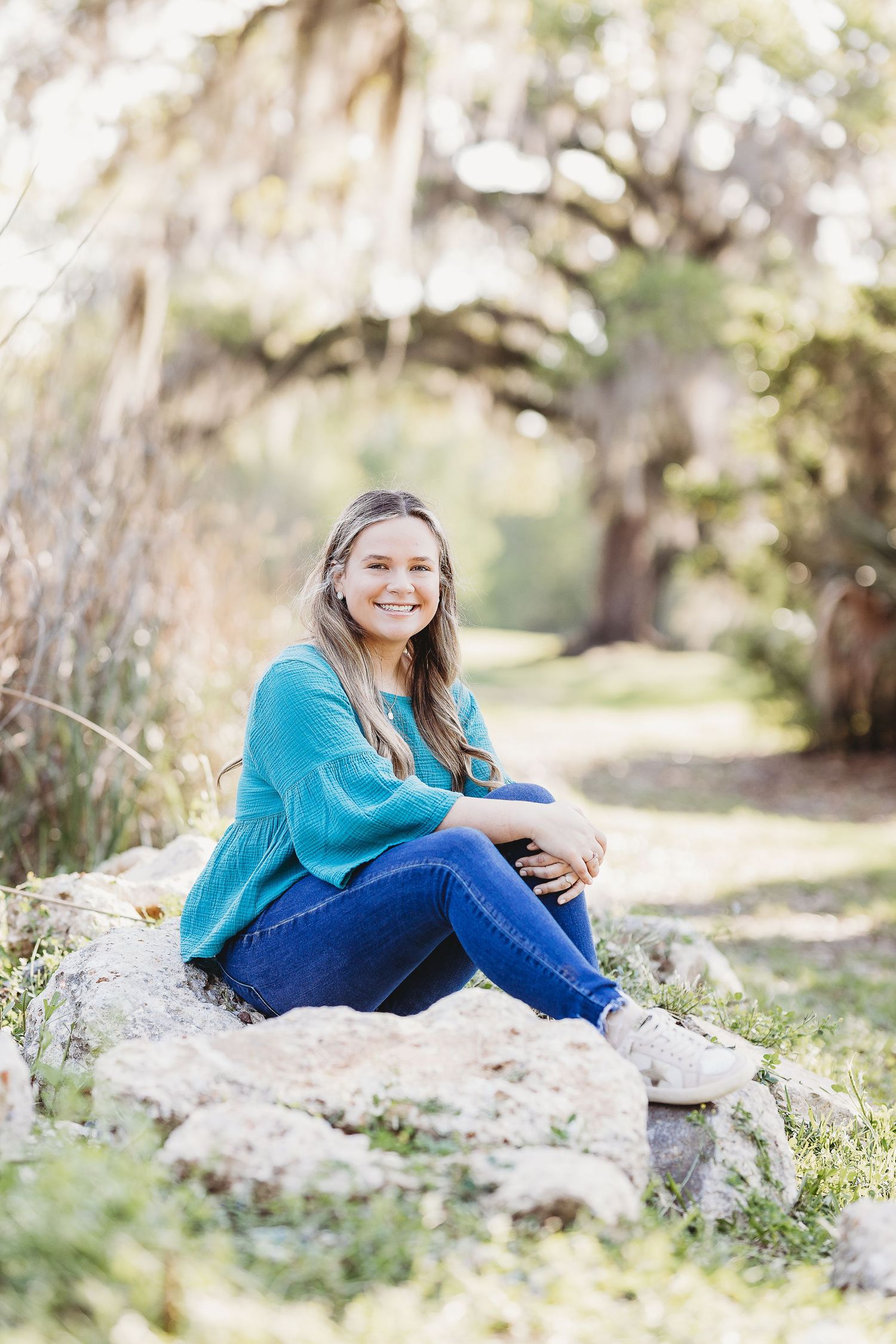 Person in turquoise sweater and jeans poses on natural rock formation in sunlit outdoor setting.