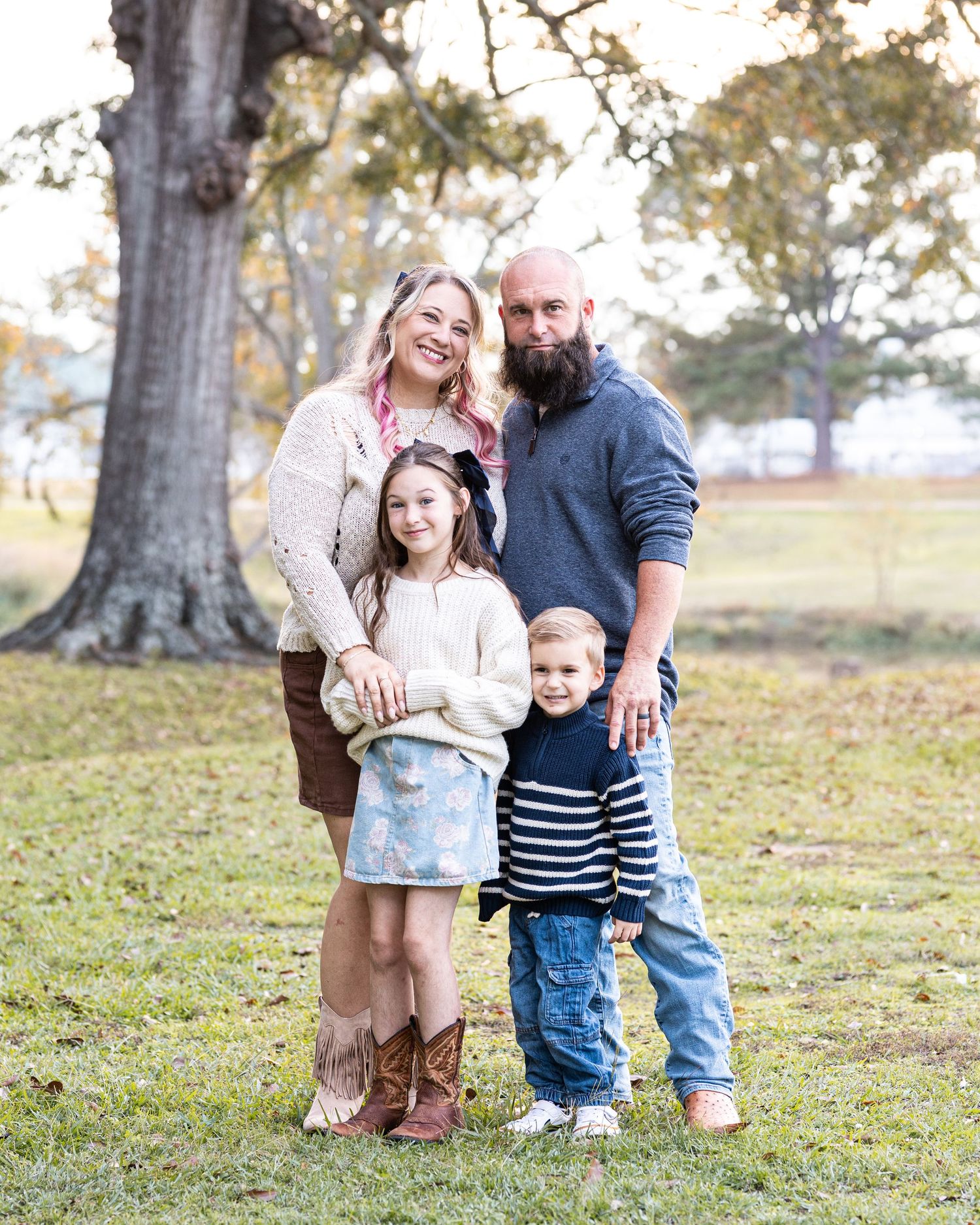 A family of four poses together outdoors in front of a large tree during autumn.