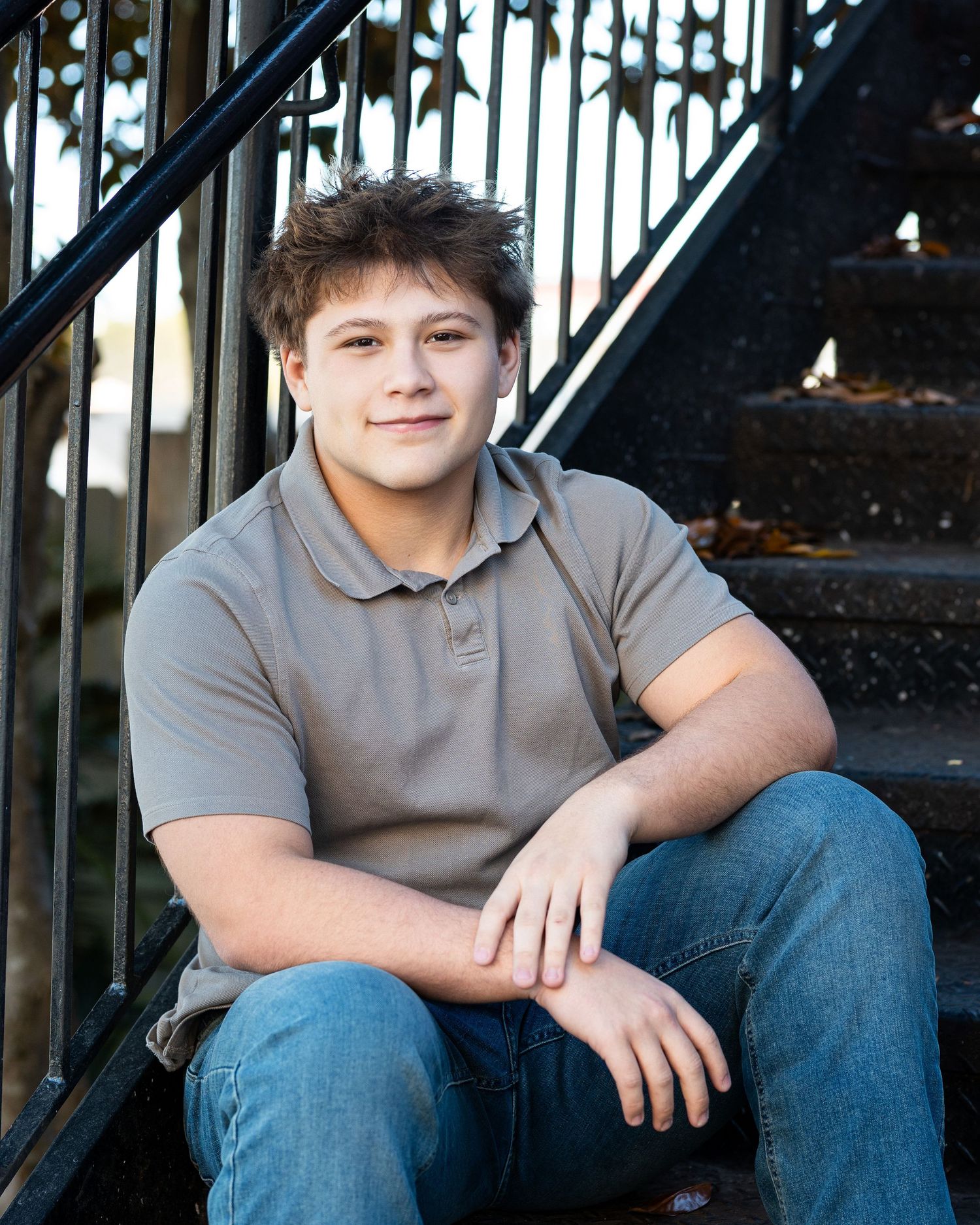 Young person in gray polo shirt and jeans sitting on dark metal stairs in casual portrait pose.