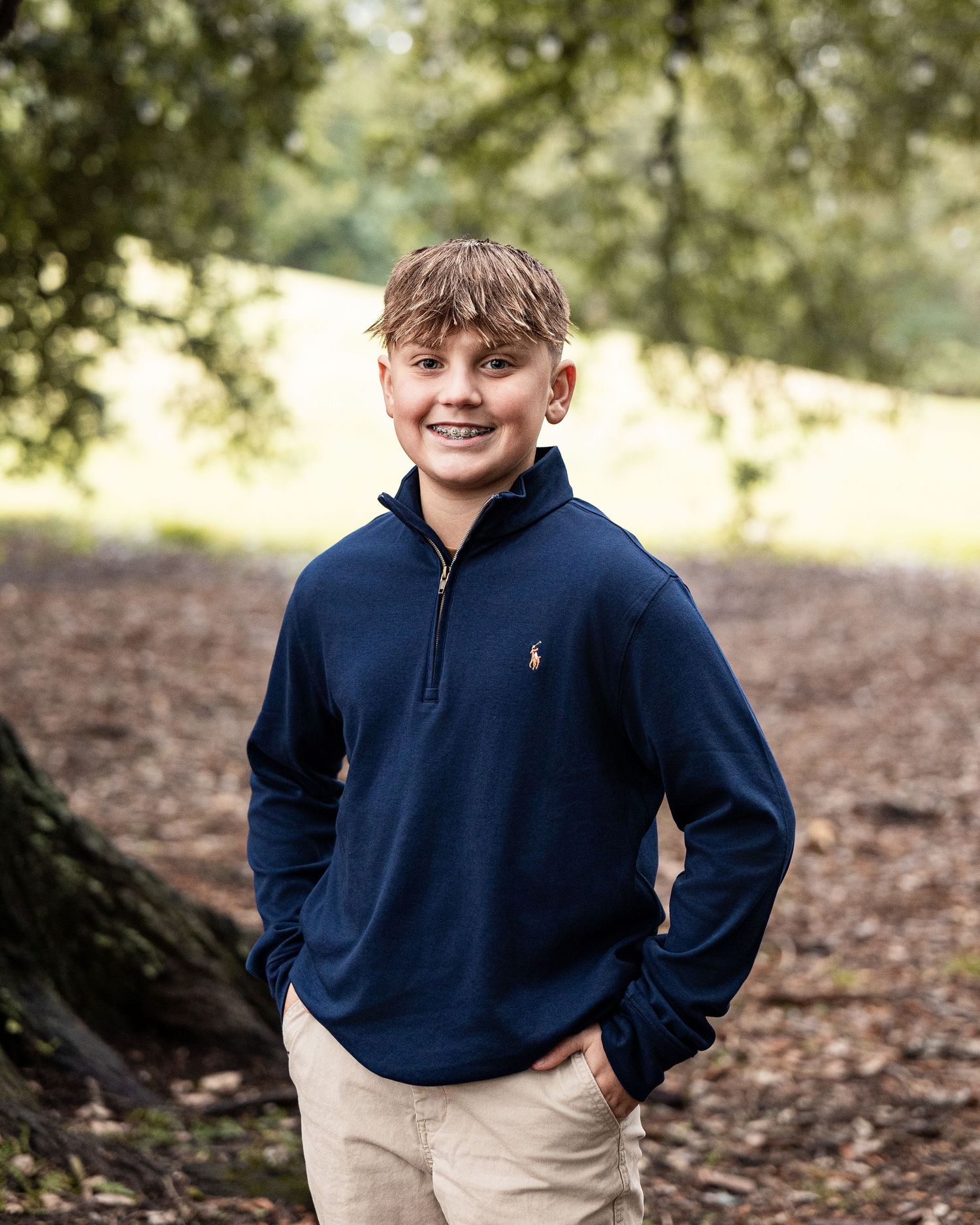 A child in a navy blue pullover and khaki pants plays outdoors on fallen autumn leaves in a natural setting.