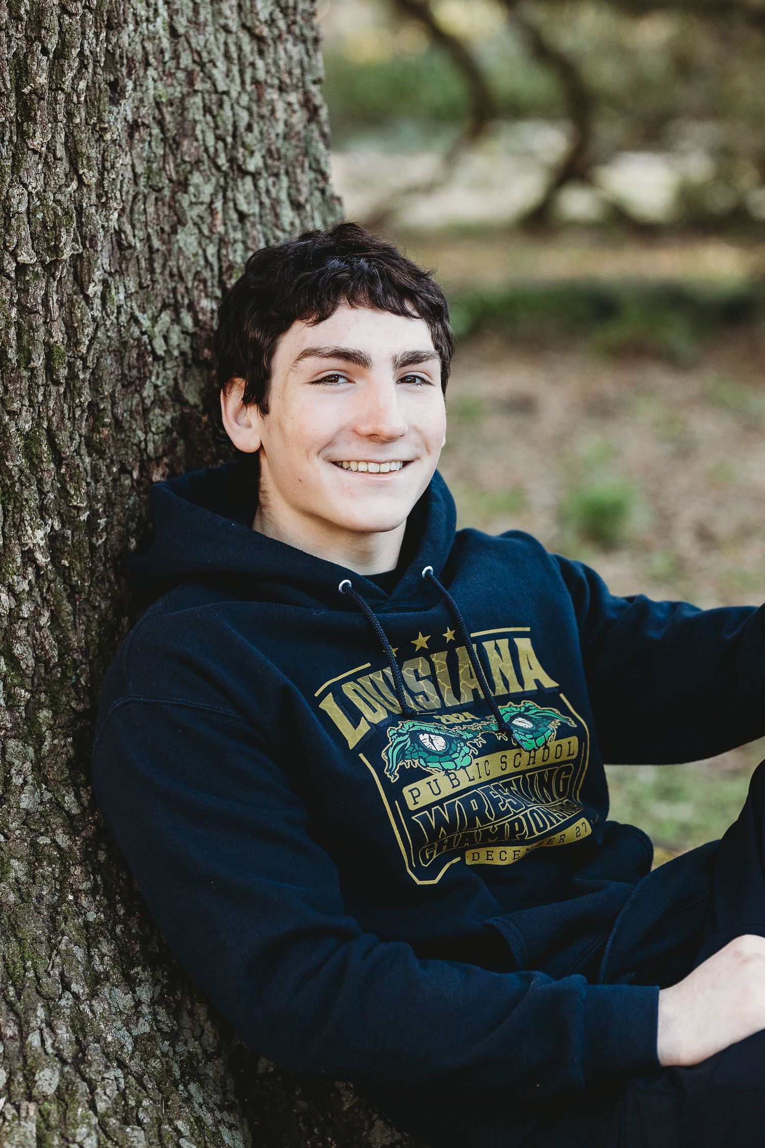 Student wearing Louisiana sweatshirt leaning against large tree trunk outdoors.