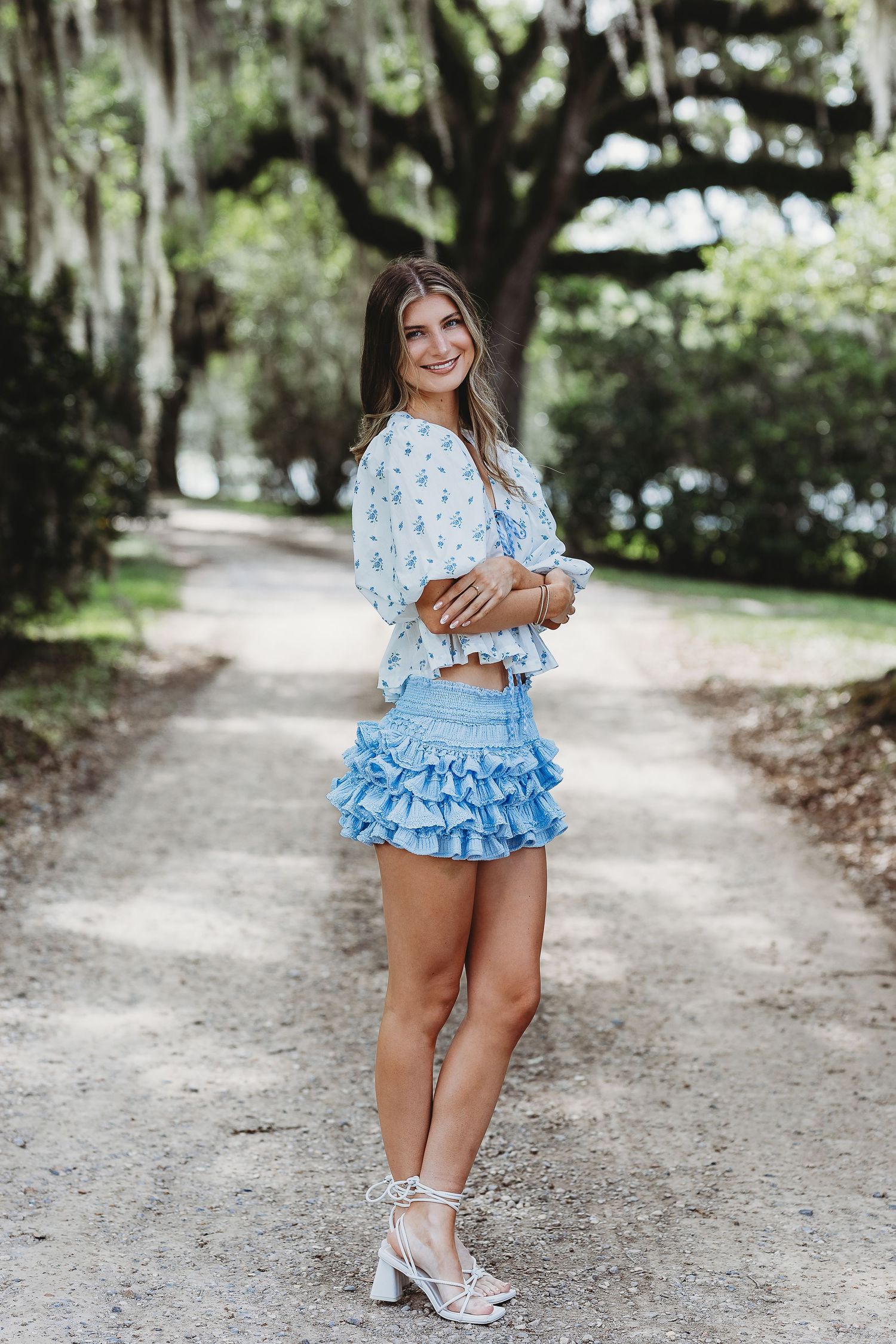 Person in light blue ruffled outfit posing on dirt path surrounded by trees.
