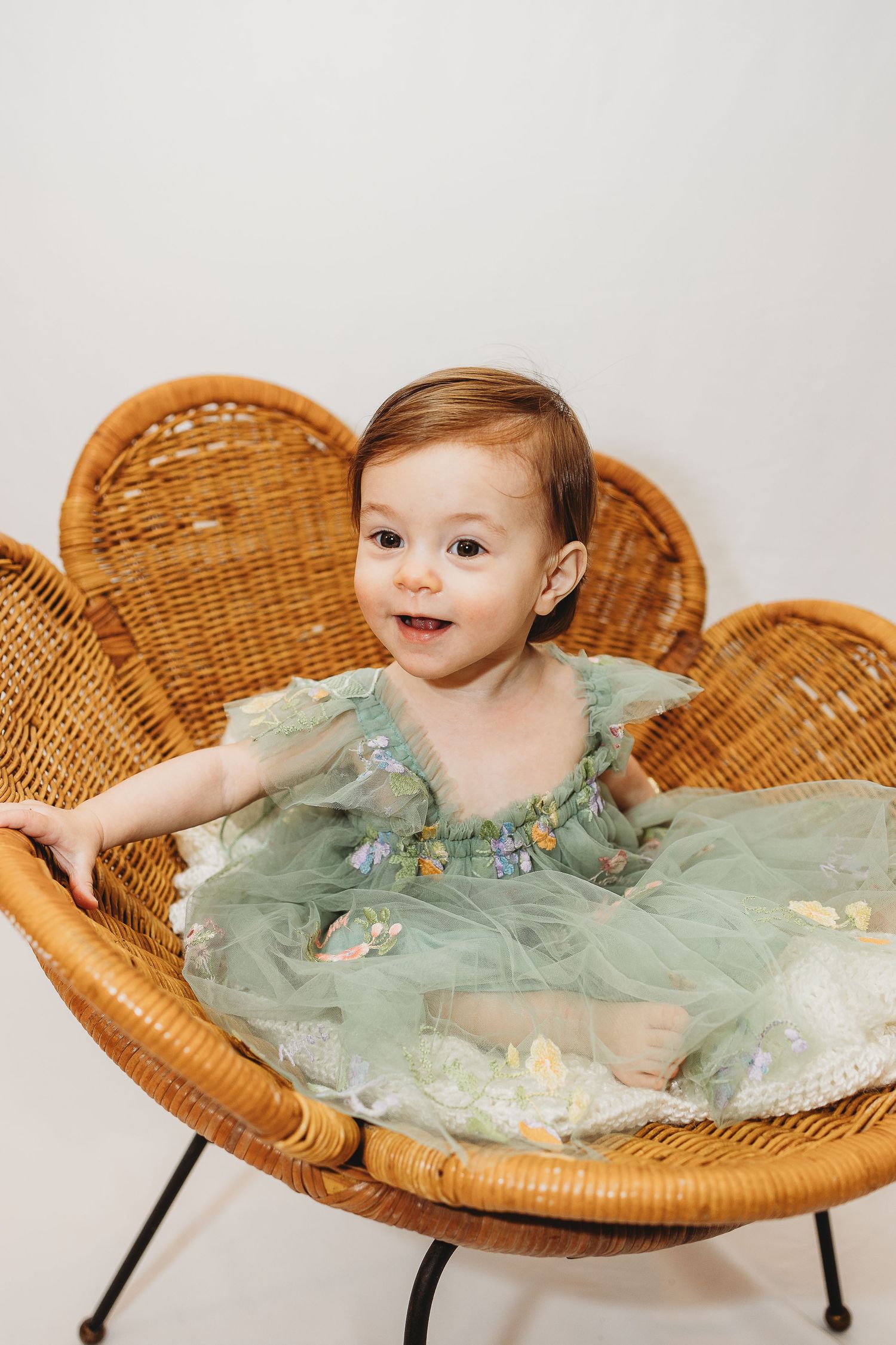 Baby in mint tulle dress sitting in wicker papasan chair against white backdrop.
