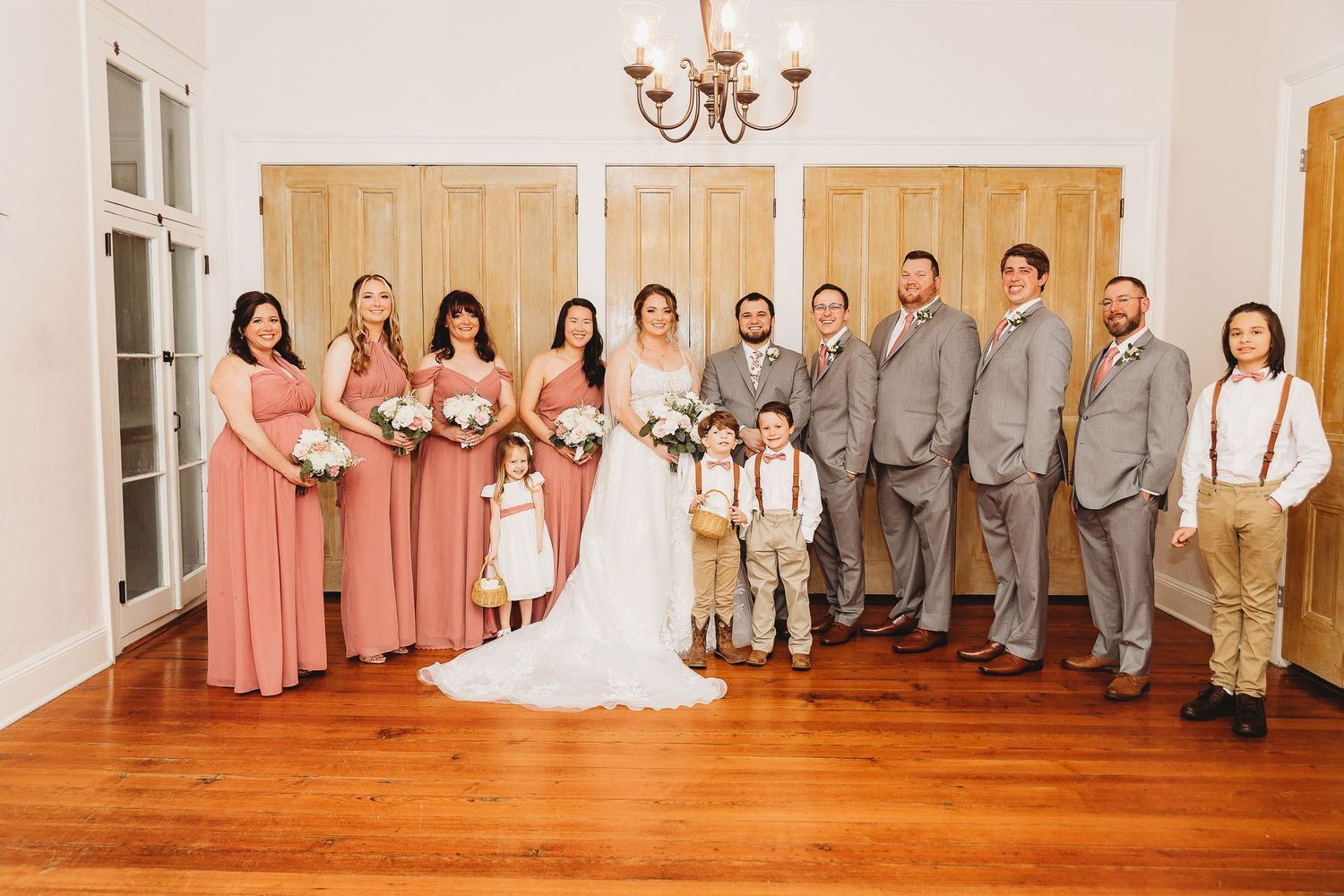 Wedding party in coral dresses and grey suits poses formally with children in a bright indoor venue.