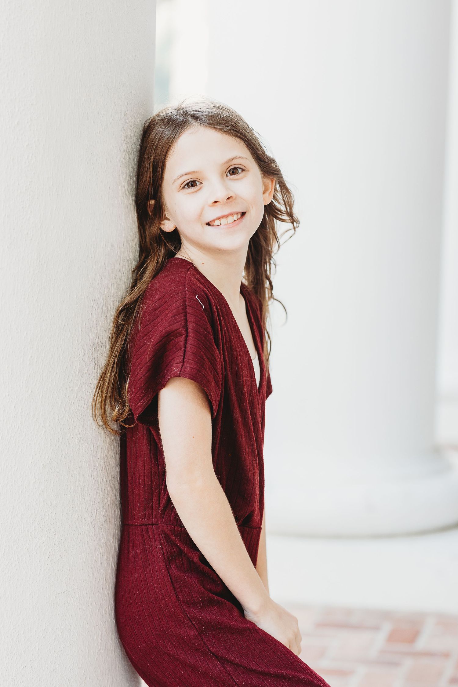 Young person in burgundy dress poses cheerfully against white column background.