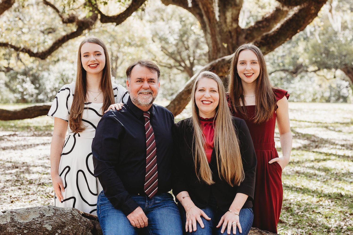 A family group poses together under large oak trees wearing coordinated burgundy and black outfits.