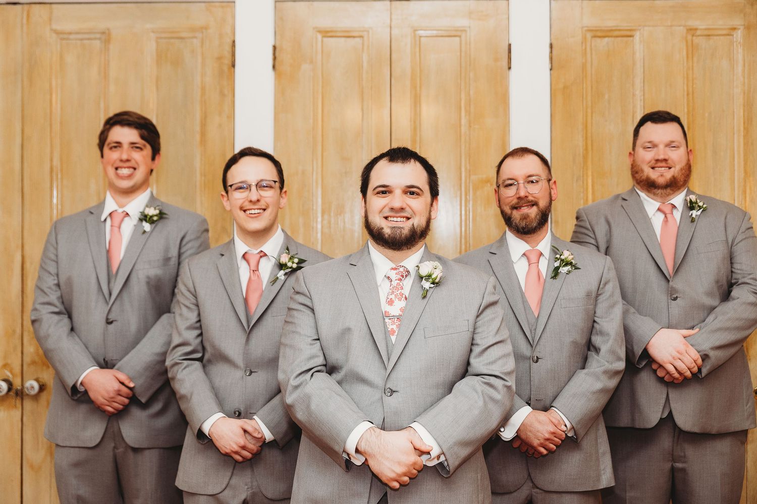 Groomsmen in matching grey suits and coral ties pose against wooden doors.