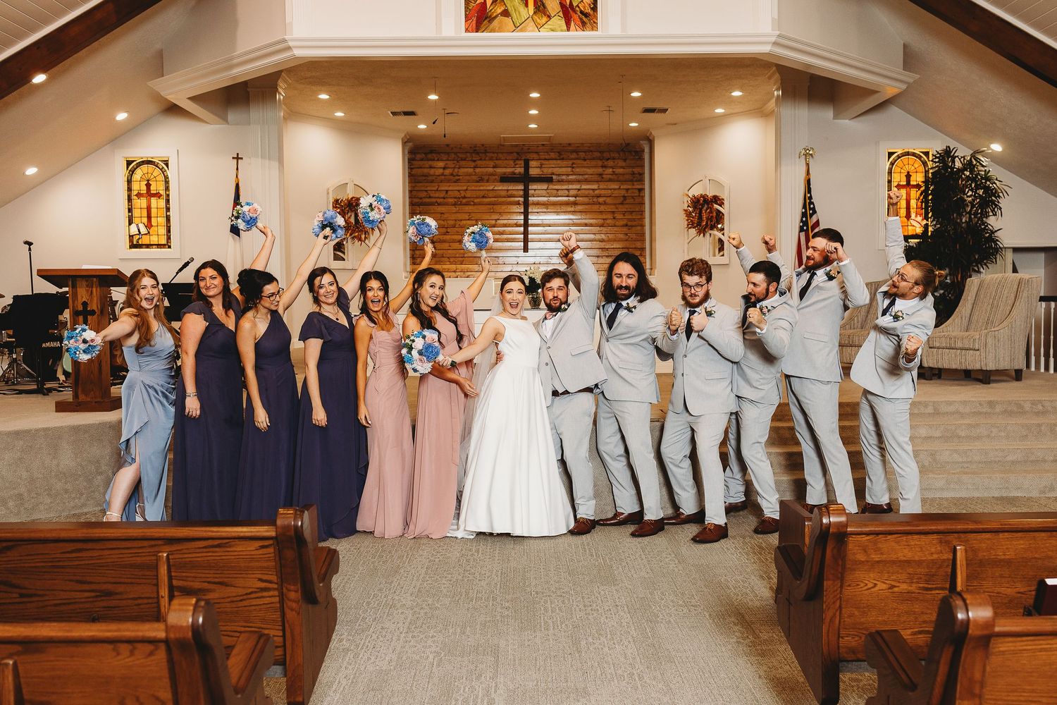 Full wedding party poses together in church sanctuary with wooden pews and warm lighting.