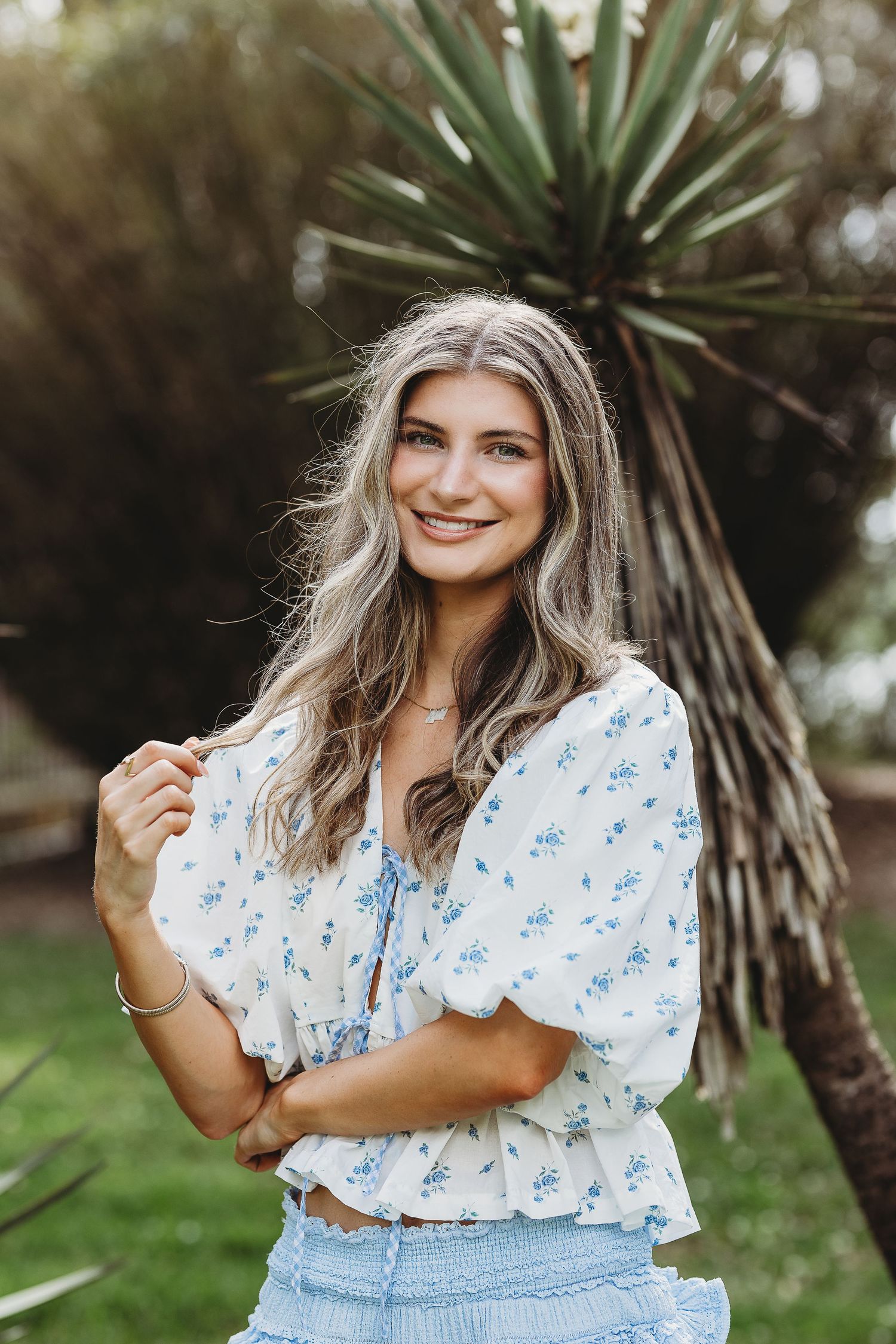 Person wearing floral print top poses in natural outdoor setting with palm plants.