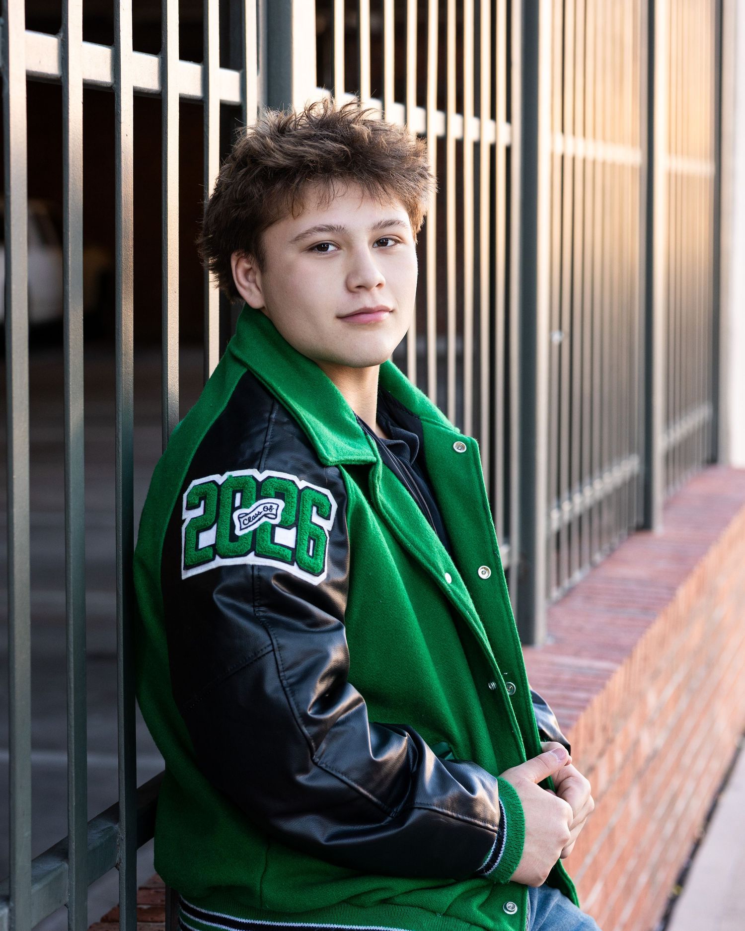 A student in a green and black varsity letterman jacket leans against metal bars in an outdoor setting.
