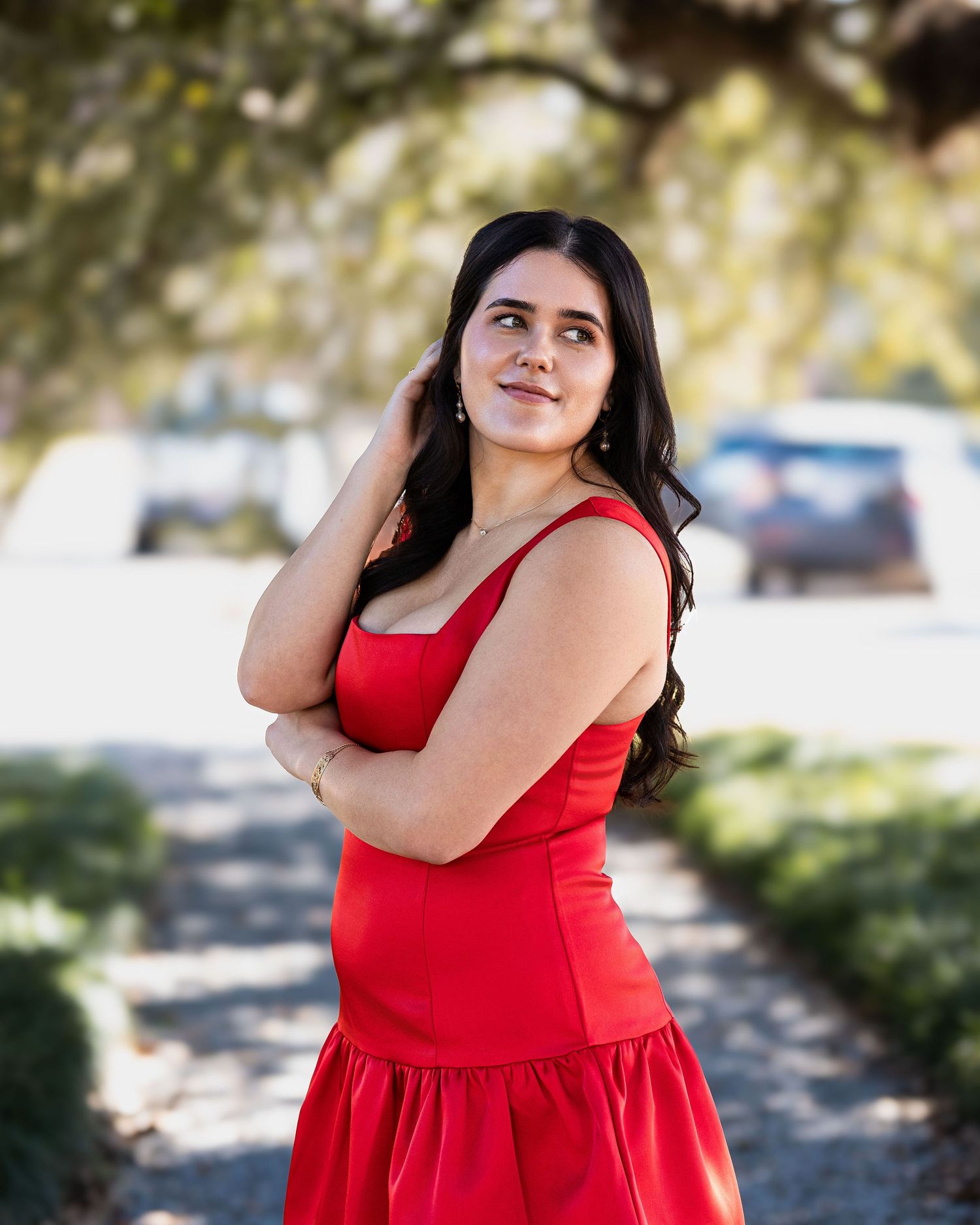 A woman in a vibrant red sundress poses outdoors on a sunlit path with trees in the background.