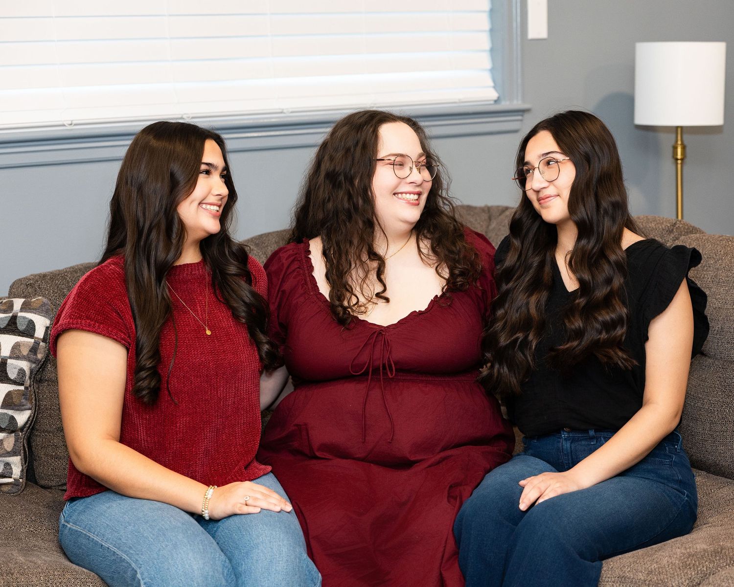 Three friends sit together on a couch, wearing casual outfits in burgundy and navy blue while sharing a joyful moment.