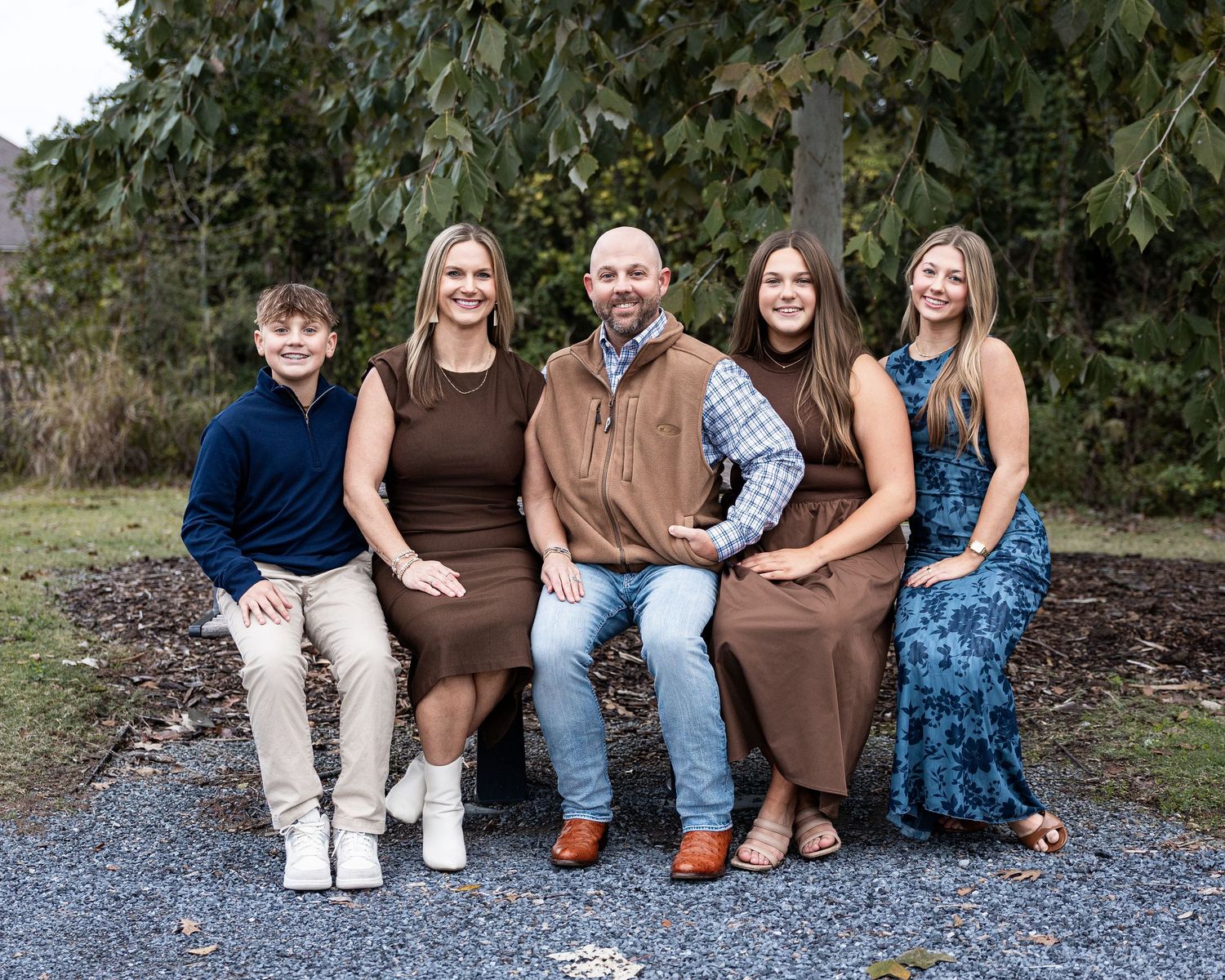 A group of people sitting together on a gravel path wearing coordinated brown and blue outfits for a family photo session.