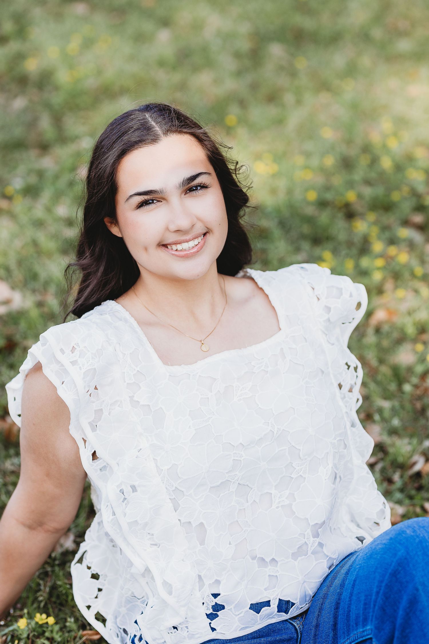 Portrait of someone in a white eyelet lace top sitting on grass with yellow wildflowers in the background.