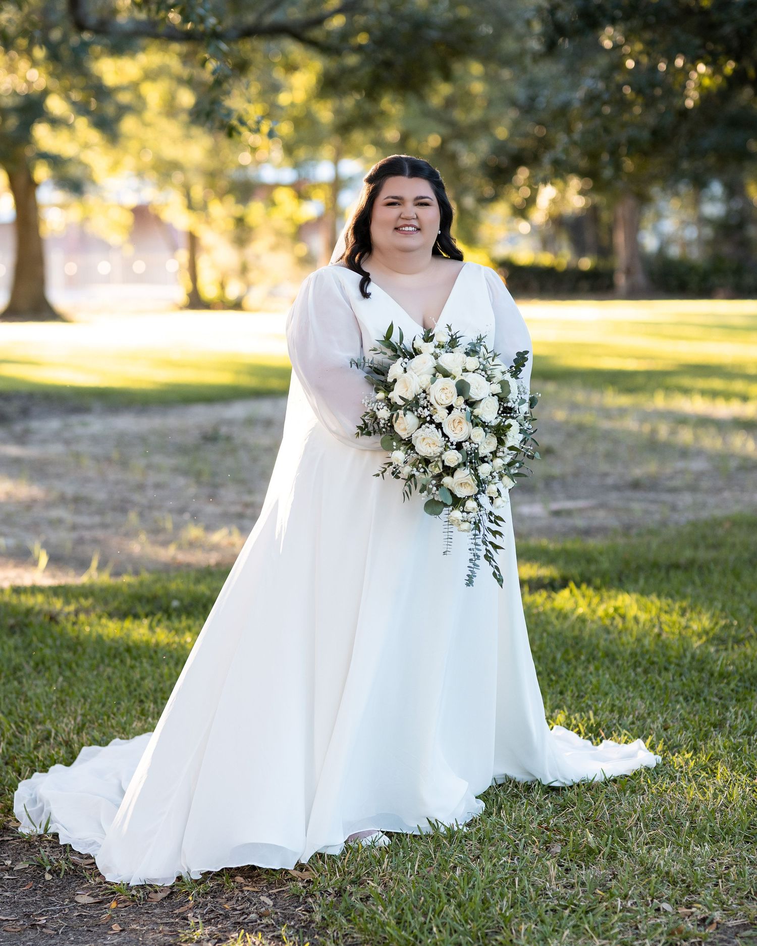 A bridal bouquet of white roses and greenery cascades down against a flowing white wedding dress in an outdoor setting.