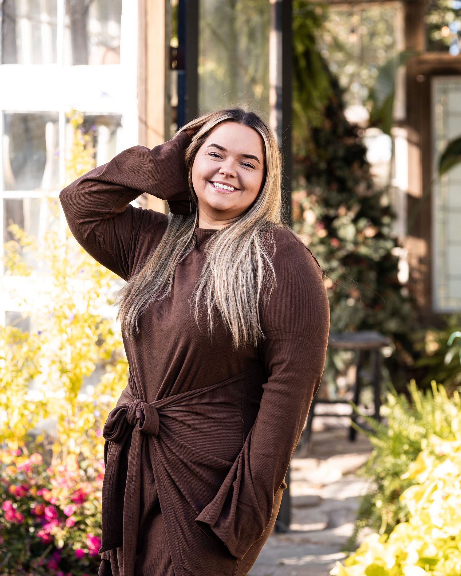 Person in brown sweater poses joyfully against outdoor backdrop with autumn foliage and flowers.