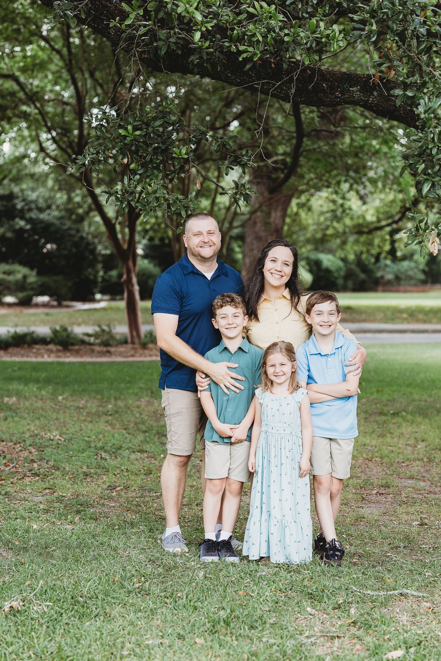 Family of five poses together under green trees in a park setting.