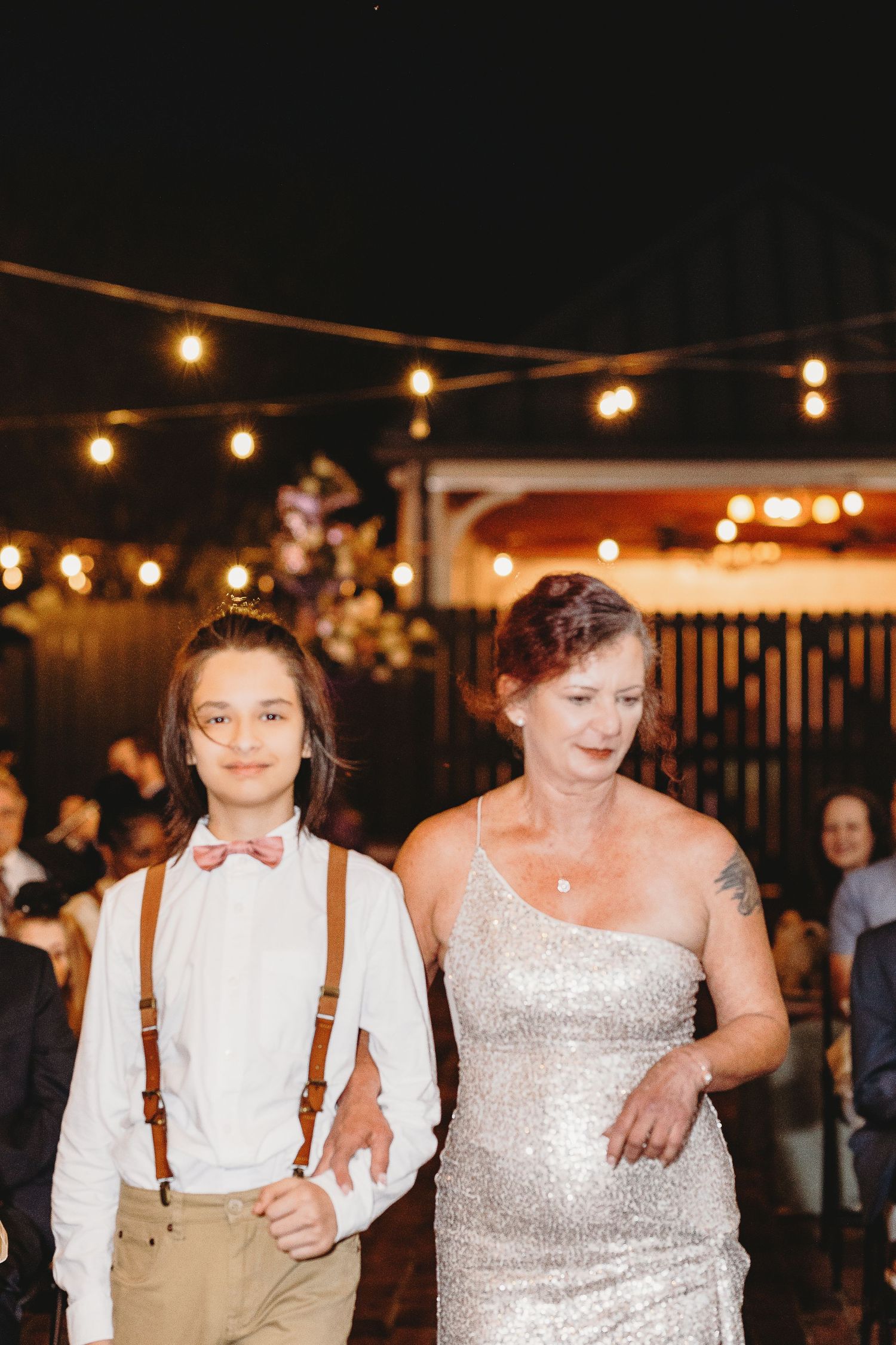 Evening wedding processional under string lights with attendants in formal attire.
