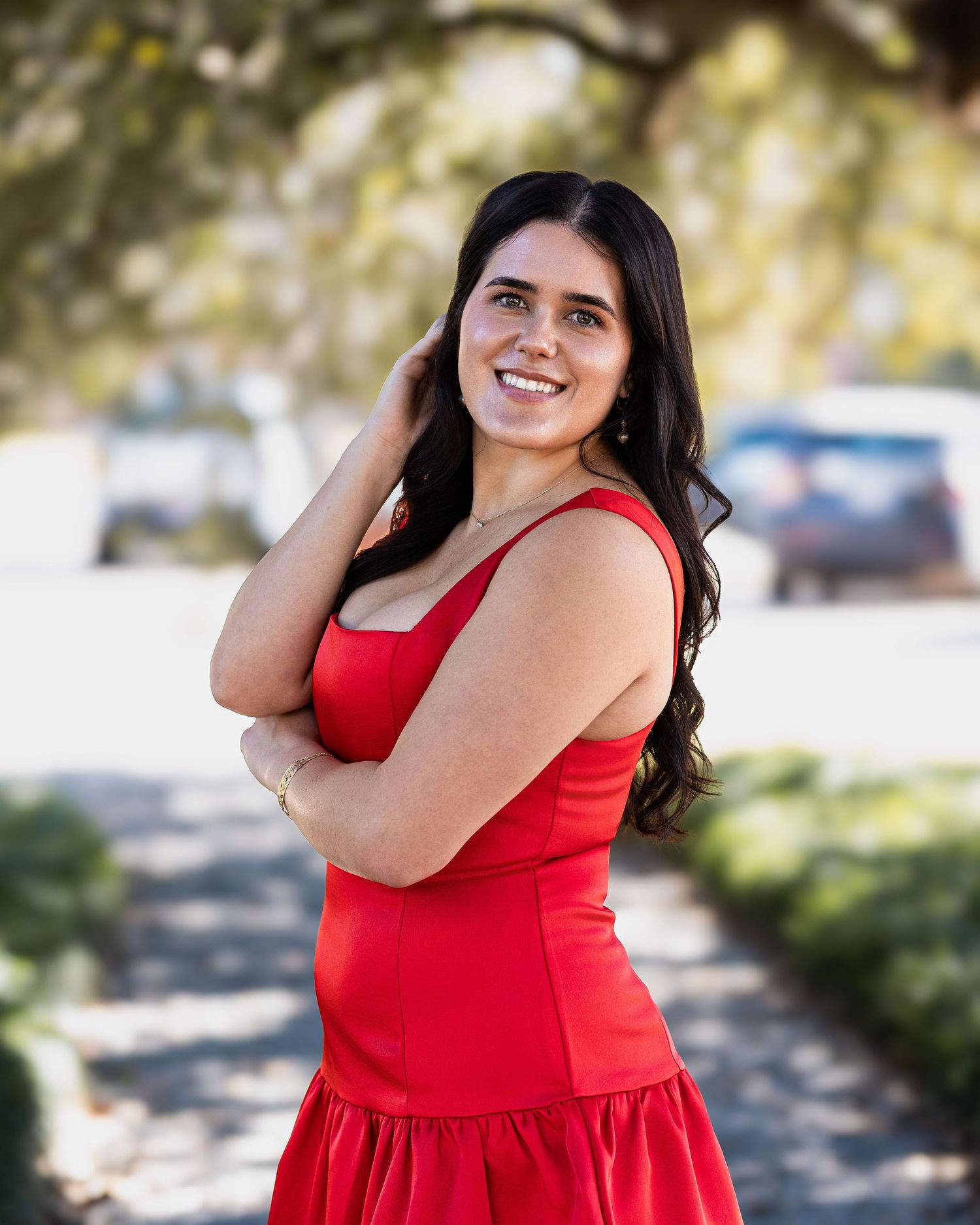 A person in a bright red sleeveless dress poses outdoors in a natural setting with sunlit trees in the background.