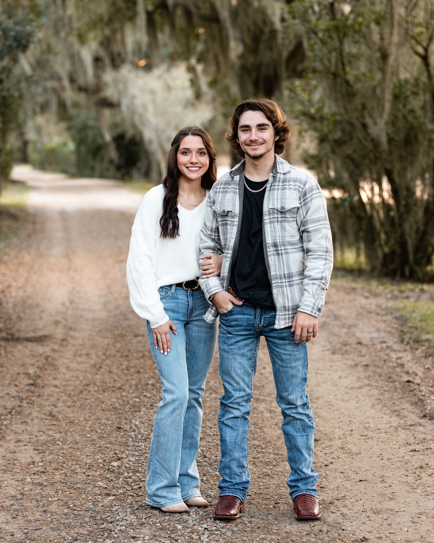 Two people wearing casual denim and plaid outfits stand together on a dirt path surrounded by trees.