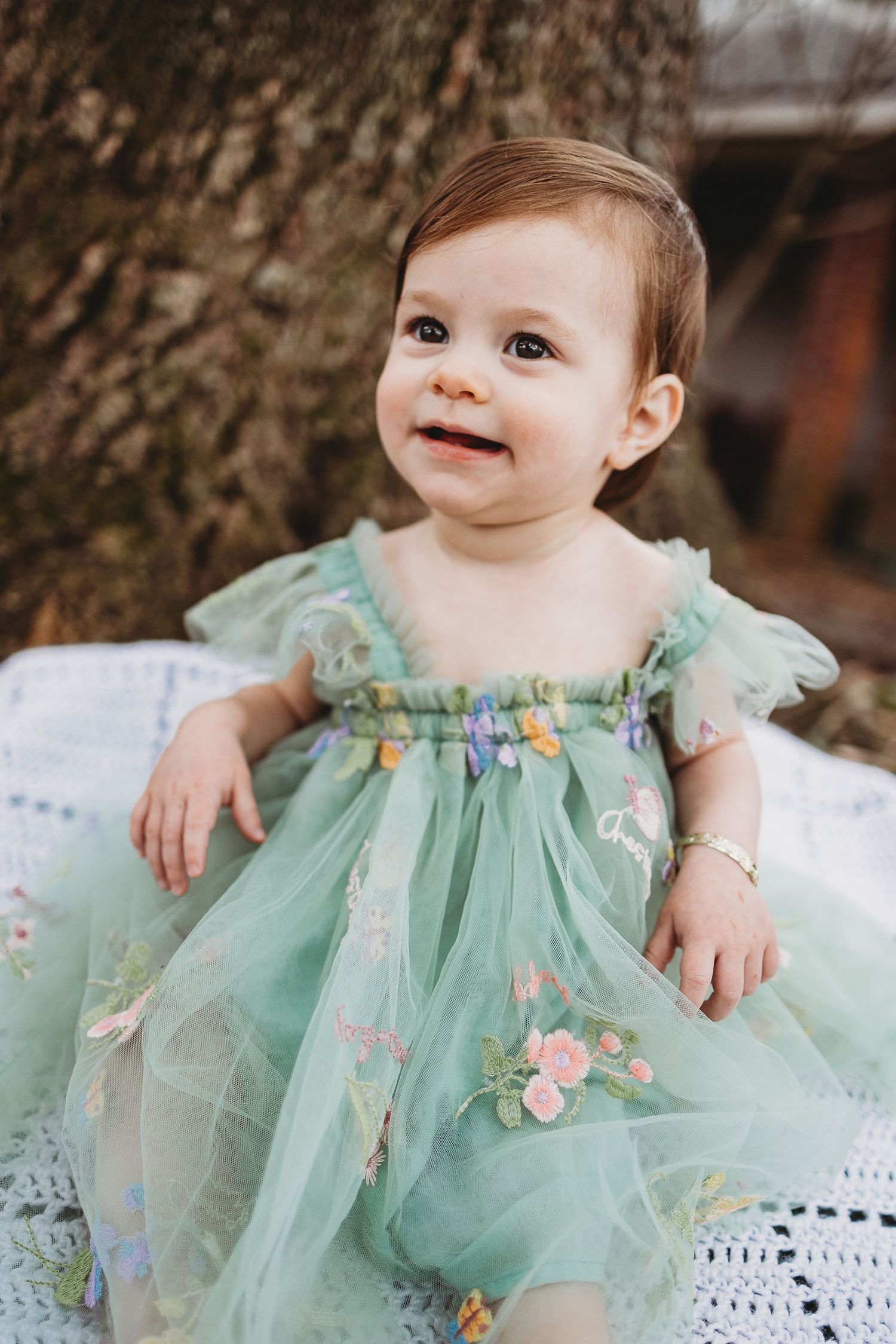 Baby wearing mint green tulle dress with floral embellishments on white backdrop.