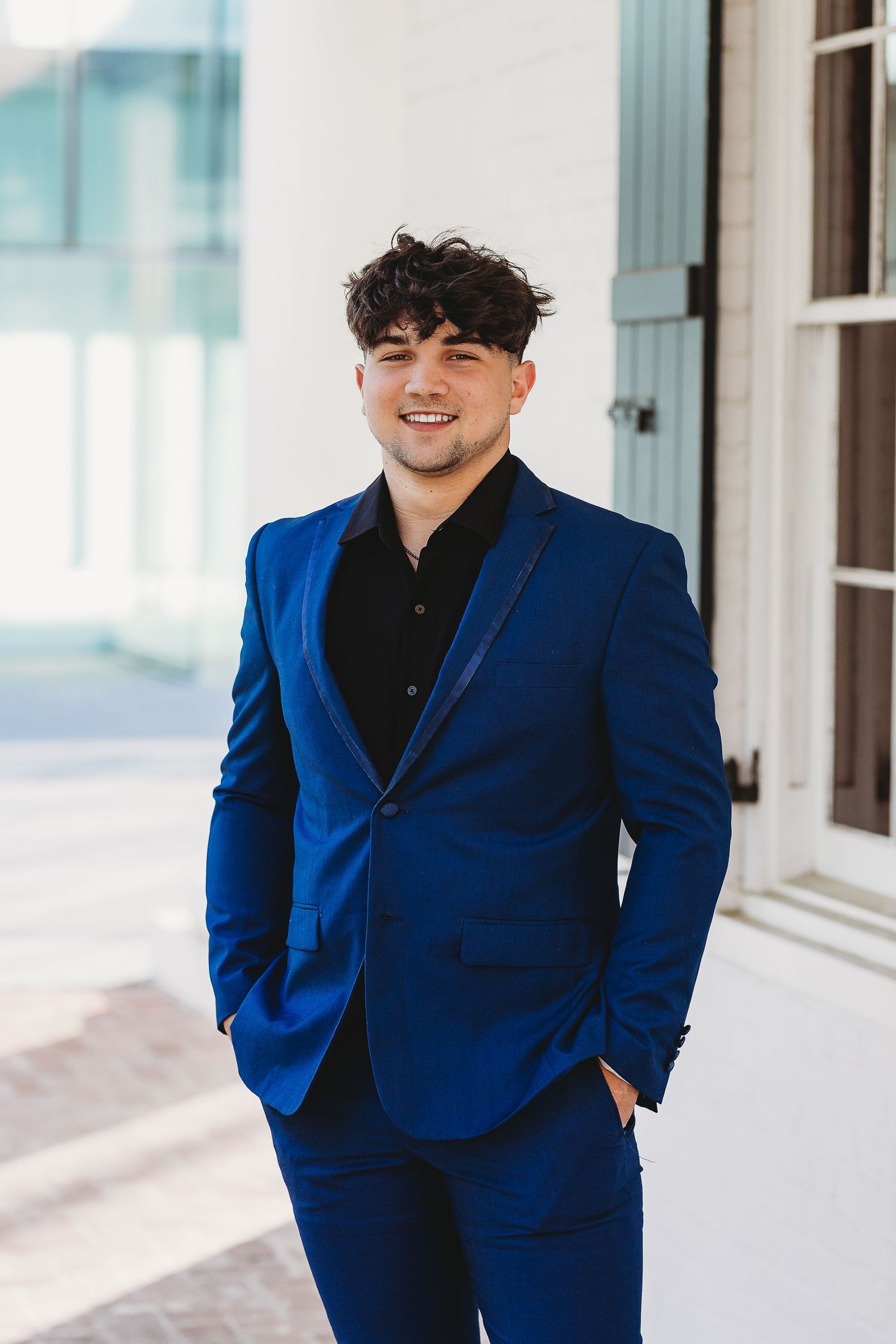Someone wearing a bright blue suit and black shirt smiles while standing by a white building.